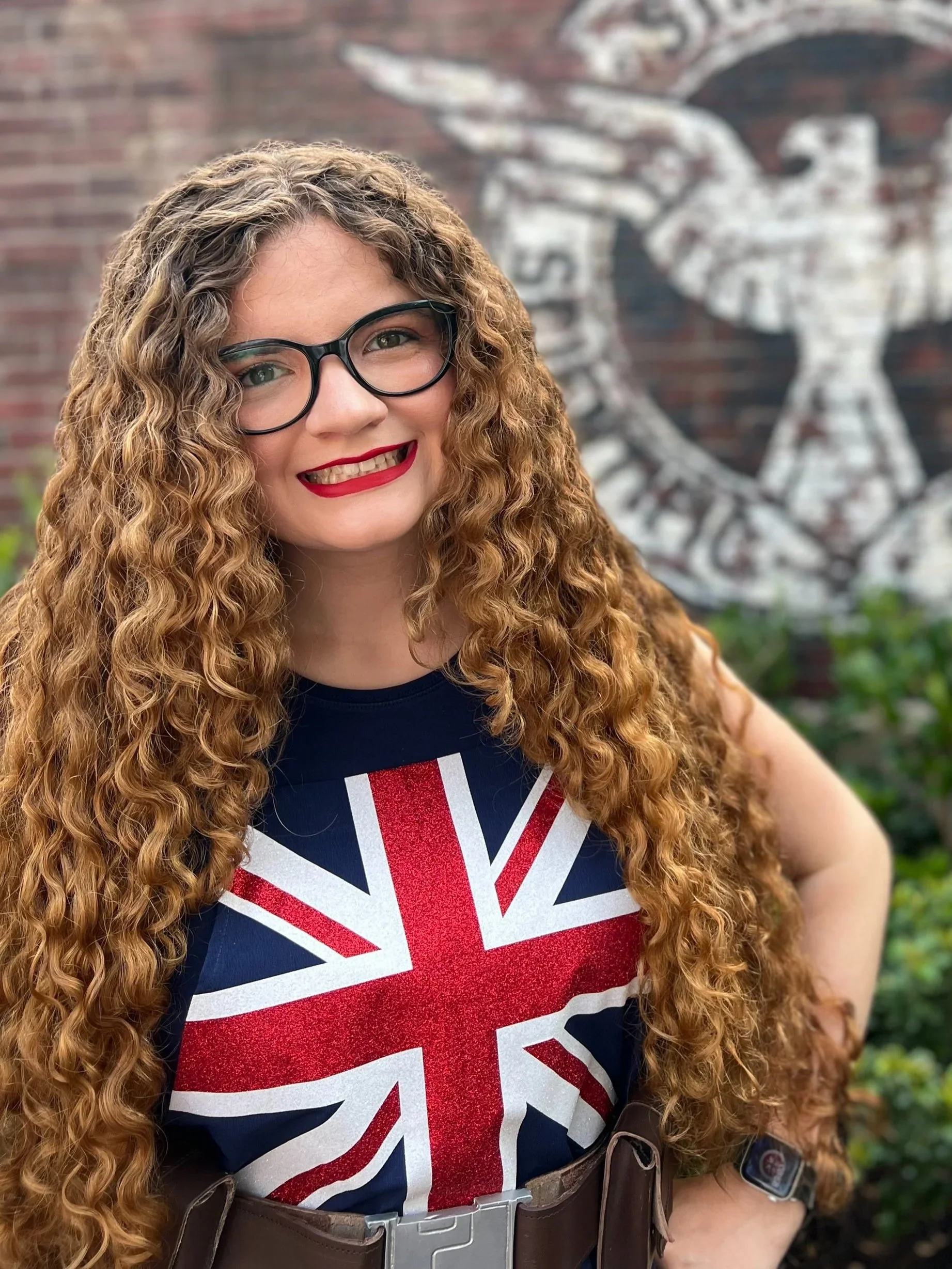 A woman with long curly red hair, glasses, and red lipstick, smiling outdoors in front of a brick wall with a large white eagle emblem painted on it. She is wearing a navy T-shirt with a Union Jack design and a watch.