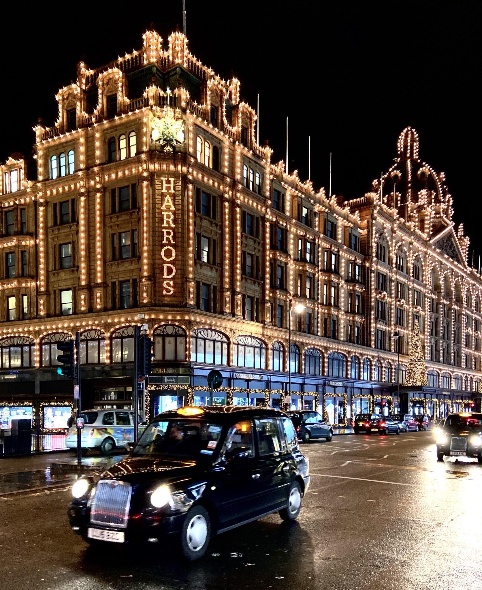 Night view of Harrods department store decorated with Christmas lights, with busy street and black taxi in the foreground.