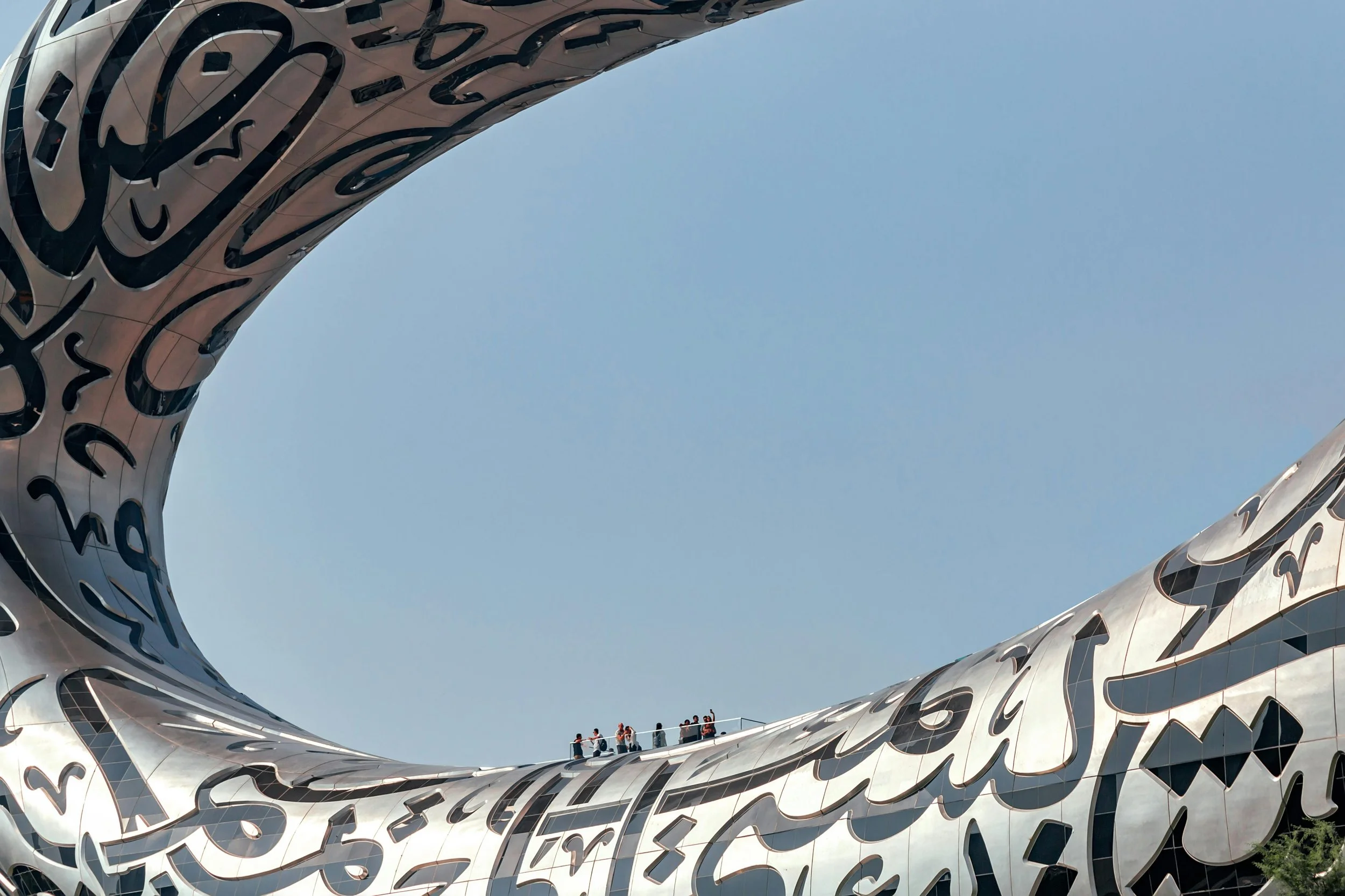 Close-up view of the unusual exterior architecture of a modern building with large Arabic calligraphy-style designs, with a partly visible group of people standing on a balcony, against a clear blue sky.