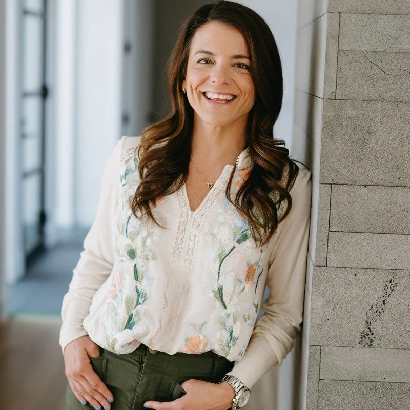 Woman with long, dark hair smiling, wearing a white embroidered blouse, standing in a modern, well-lit indoor space next to a gray stone wall.