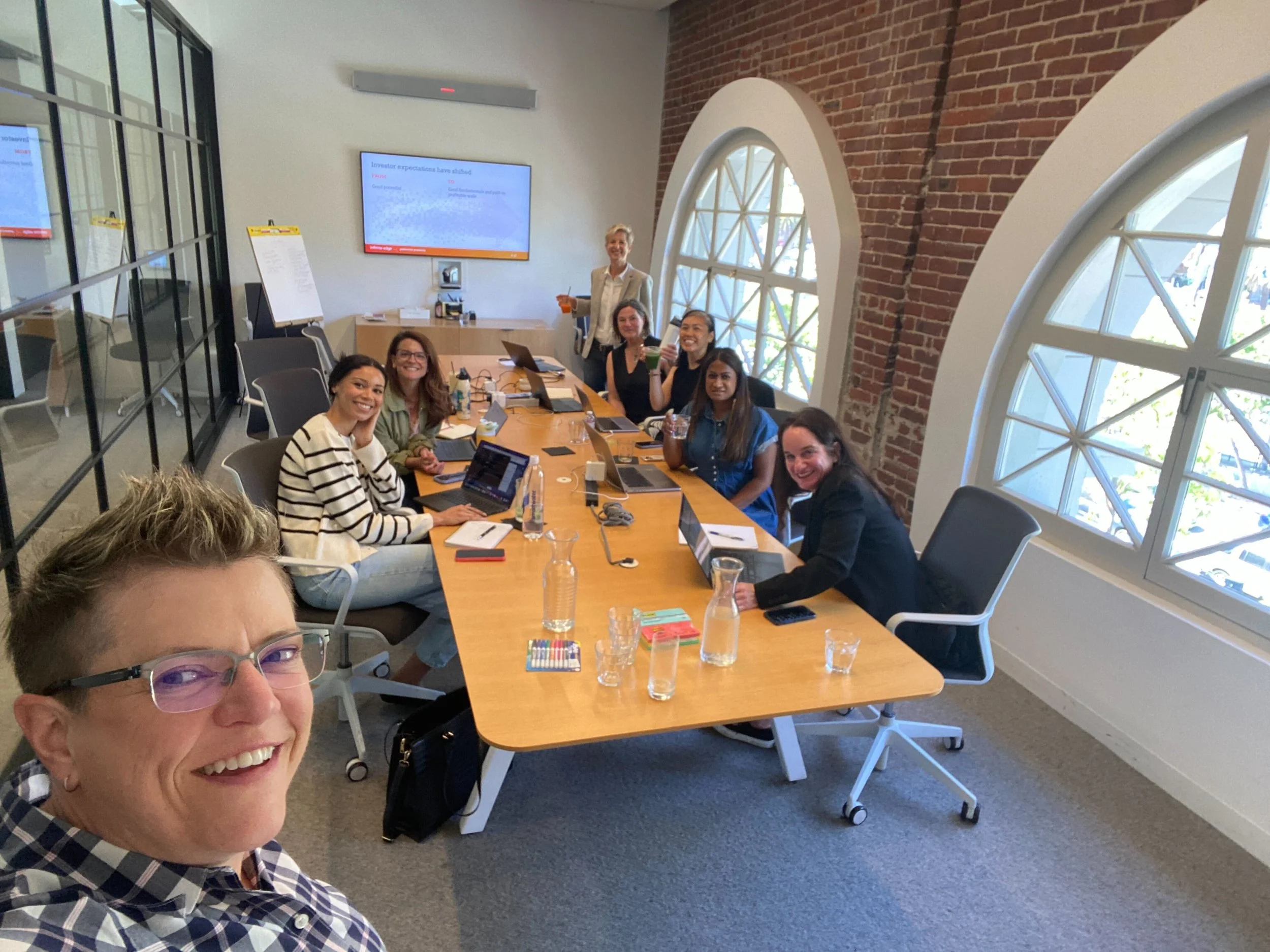 A group of people in a conference room smiling and taking a selfie at a meeting table with laptops, notepads, and drinks, with a woman at the back standing and holding a drink. Large arched windows on the right side let in natural light.