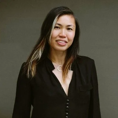 A woman with long dark hair smiling and wearing a black blazer against a neutral background.