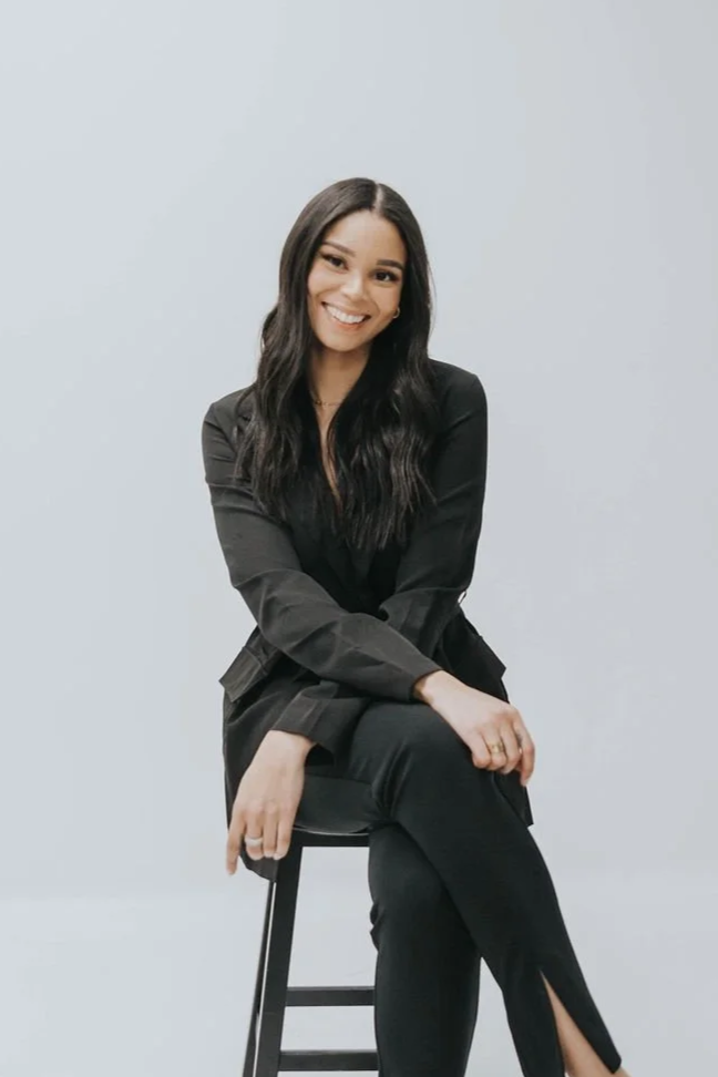 A woman with long dark hair wearing a black blazer and pants, sitting on a stool with a neutral gray background.