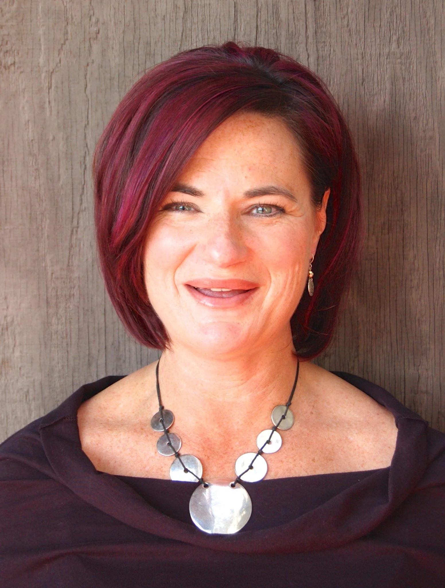 A woman with short burgundy hair, bright blue eyes, and freckles, smiling in front of a wooden background, wearing a black top and a large silver necklace.