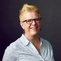 A woman with short blonde hair, wearing glasses and a white shirt with small polka dots, smiling against a dark background.