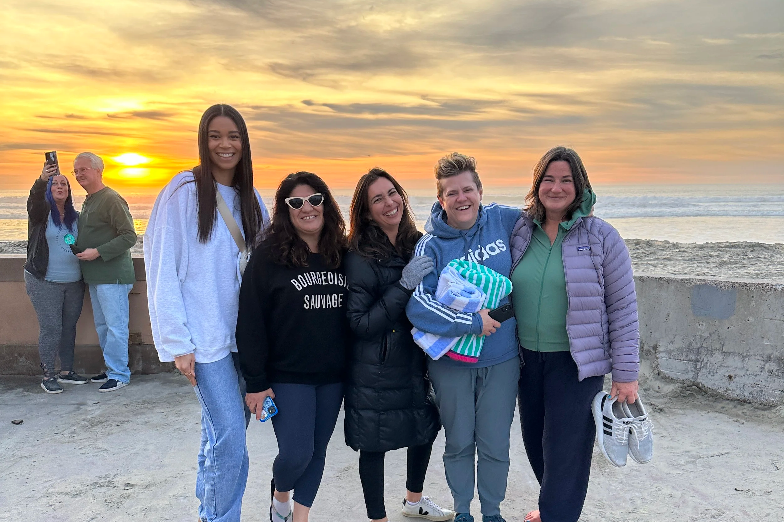 Five women standing on a beach at sunset, smiling for a group photo, with two people taking a picture in the background.