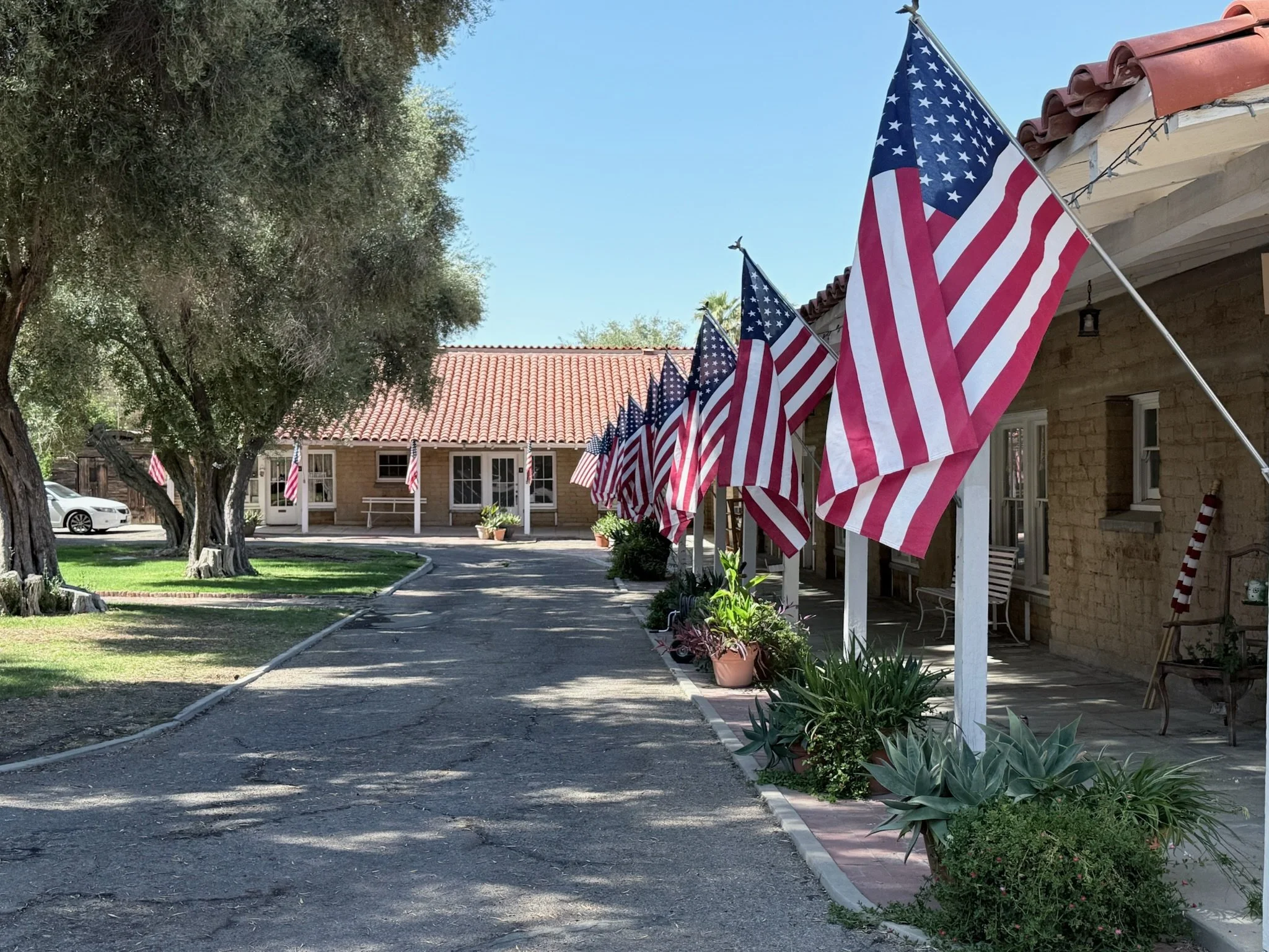 American flags lining the porch of a building with a tiled roof, with a tree and parked car in the background on a clear day.