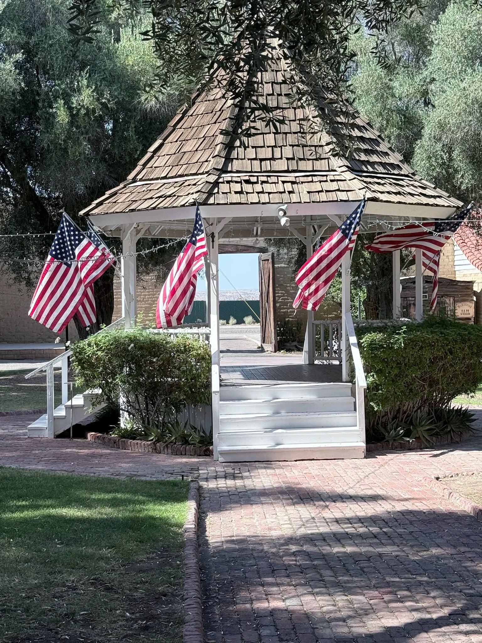 A small white gazebo decorated with American flags, situated on a brick pathway in a garden with bushes and trees.