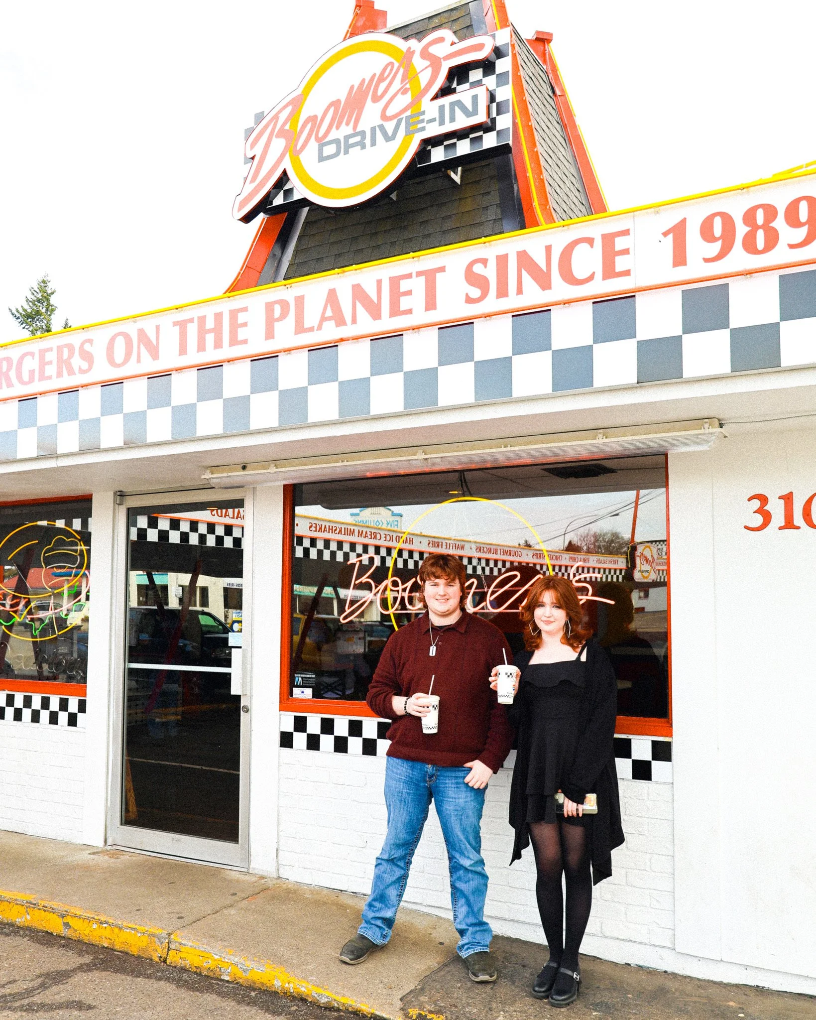 Two young people standing outside a Boomers Drive-In getting burgers. They are holding milkshakes and smiling. The restaurant has checkered black and white patterns and neon signs.
