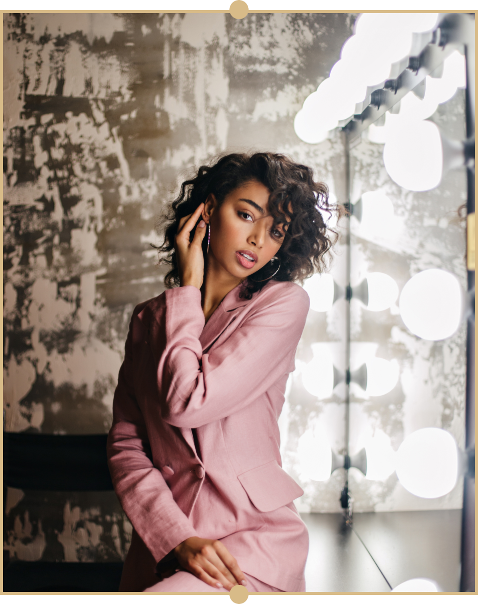A woman with curly dark hair and hoop earrings, wearing a pink blazer, posing in front of a mirror with bright round lights.