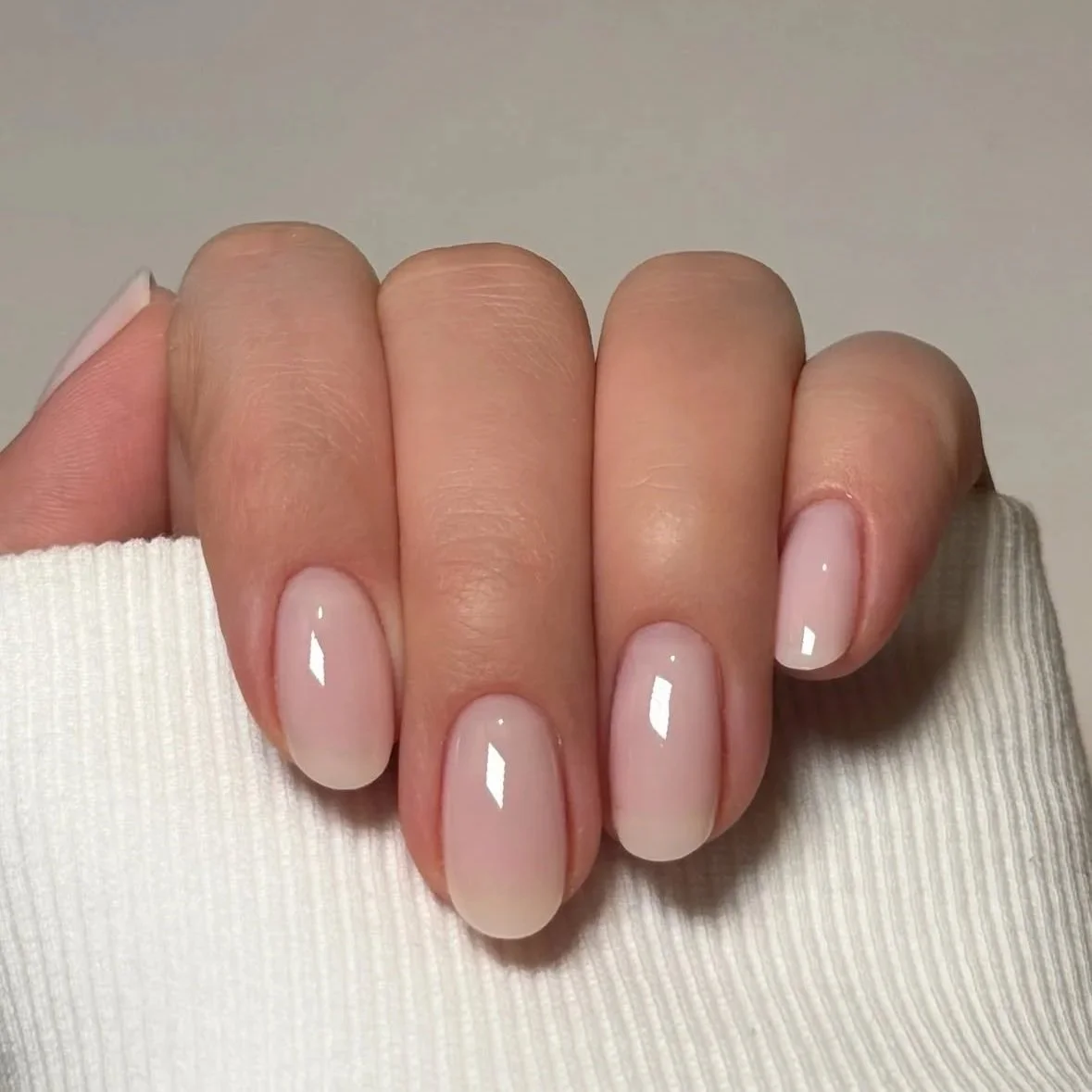 Close-up of a hand with manicured nails painted in a shiny, nude color, wearing a white ribbed sweater