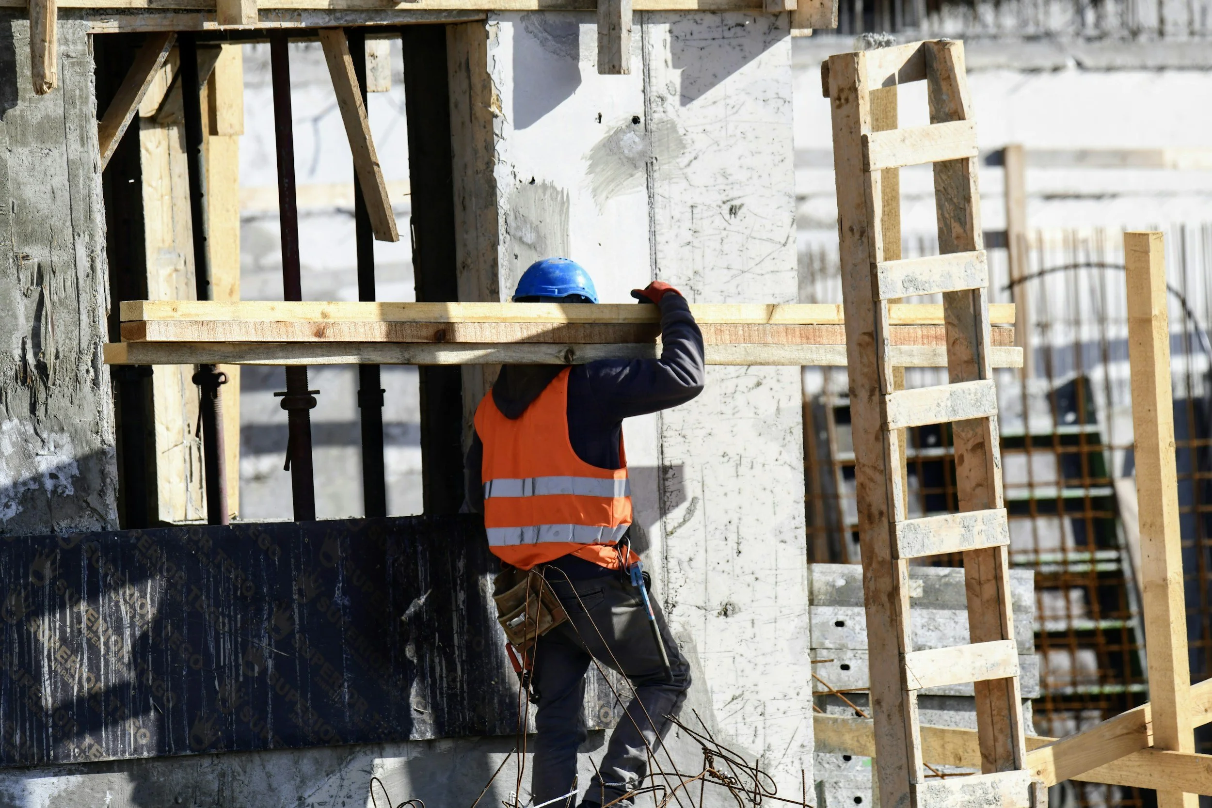 Construction worker wearing a blue helmet, orange safety vest, and gloves on a building site, carrying a wooden plank, with a ladder leaning against the structure.