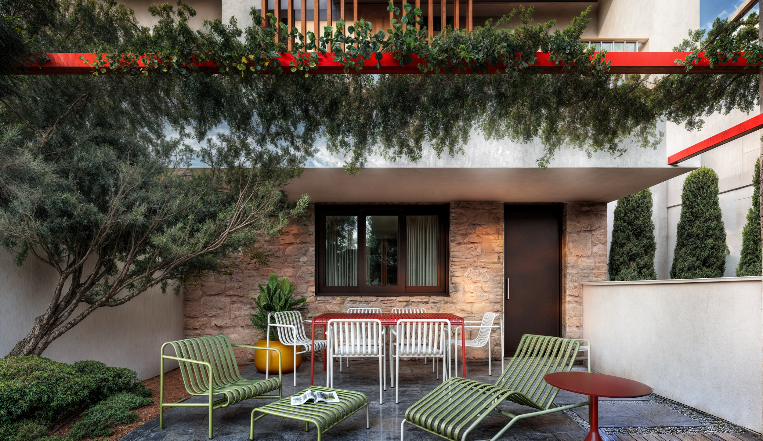 Outdoor patio with modern furniture including green and white lounge chairs, a red table, and white chairs with a red table. A potted plant is beside a window on a stone wall. There is greenery, including trees and tall bushes, around the patio area.