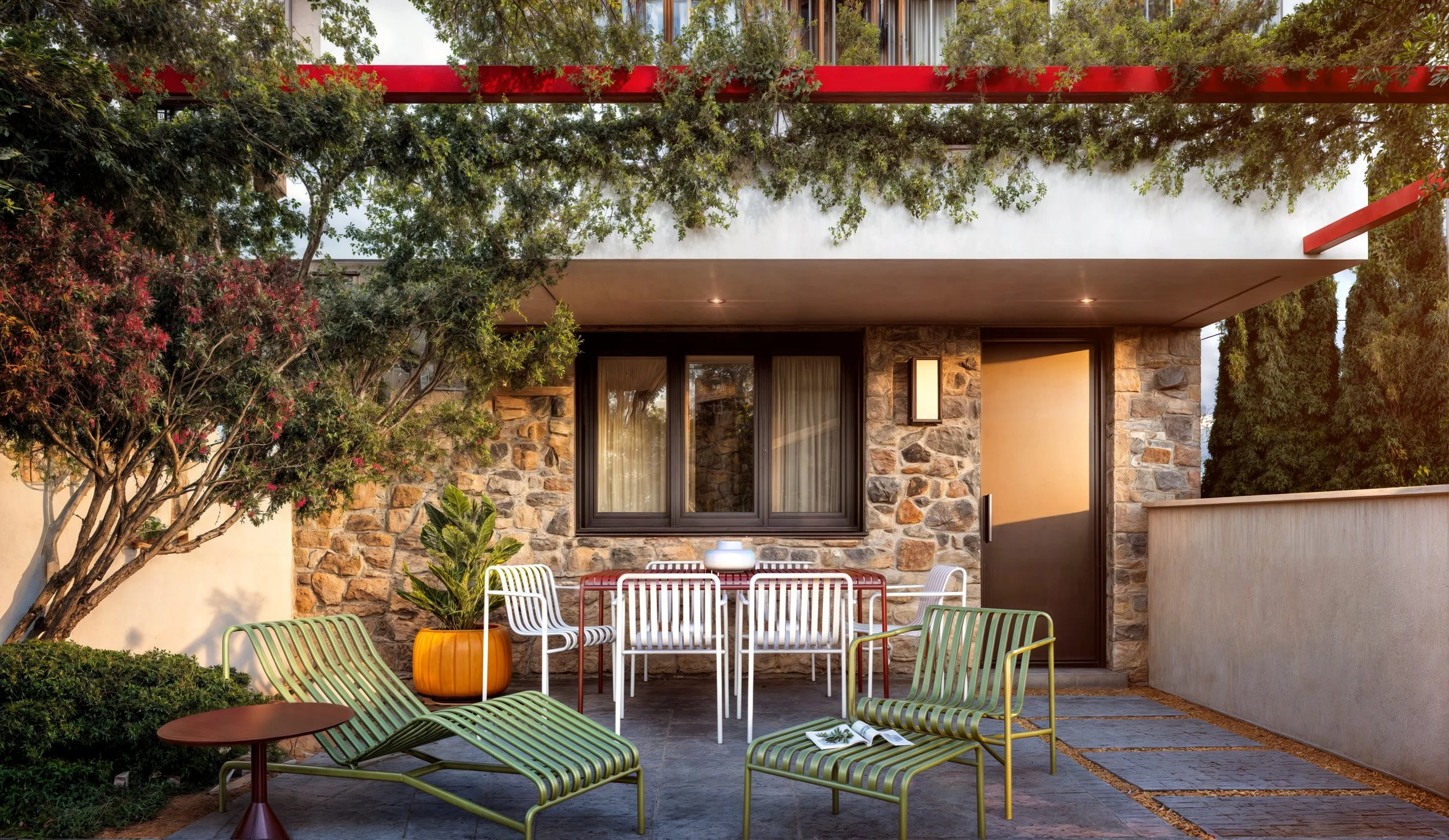 Outdoor patio with green and white striped lounge chairs, a small round table, a white dining table with multiple white chairs, potted plants, trees, and a stone wall background.