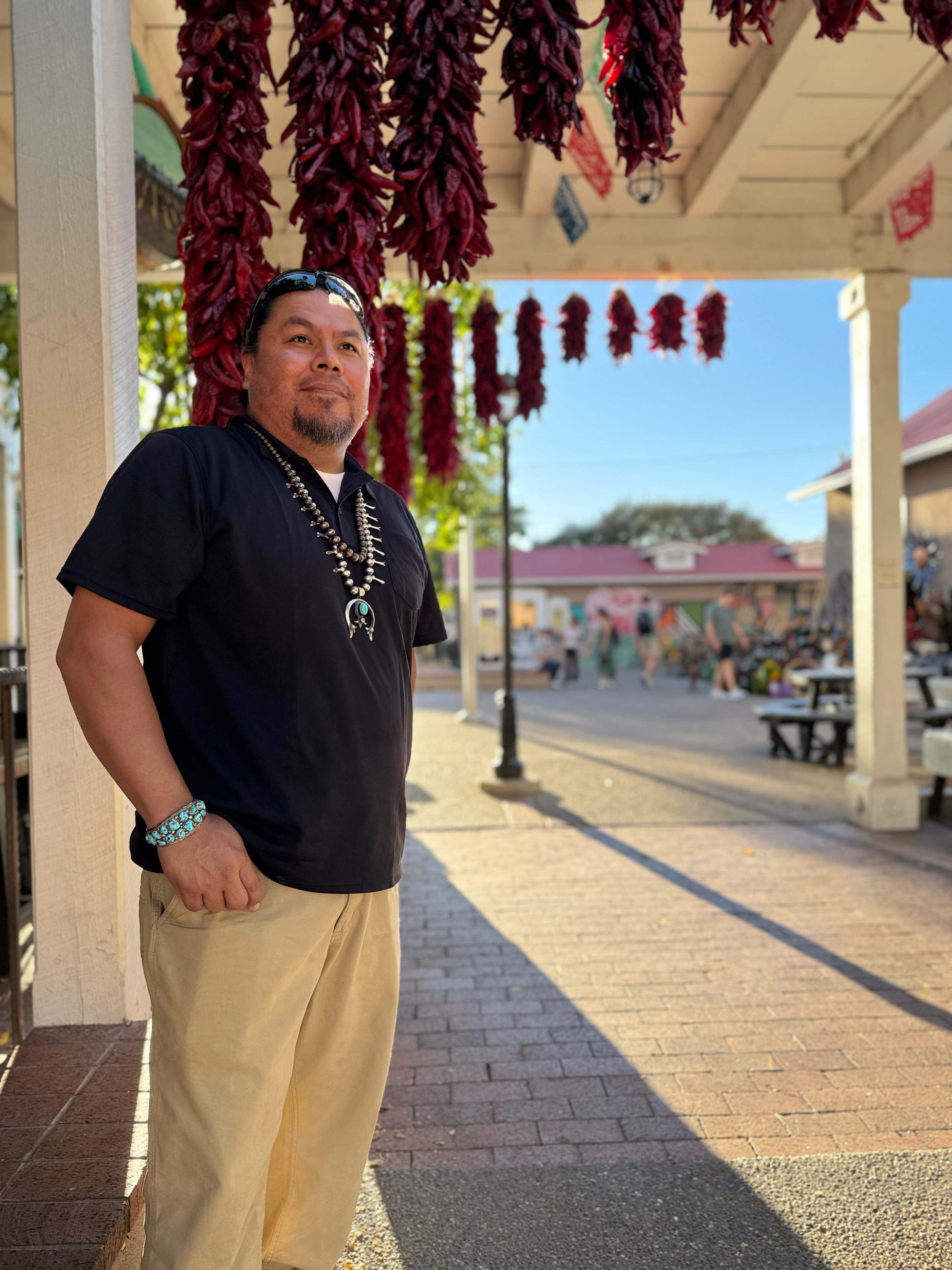 A man standing near a wooden structure with hanging red chili peppers and colorful banners in the background on a sunny day.