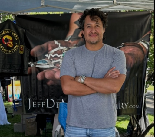 A man stands in front of a booth with a Jeff DeMent banner, smiling with arms crossed, outdoors on a sunny day.
