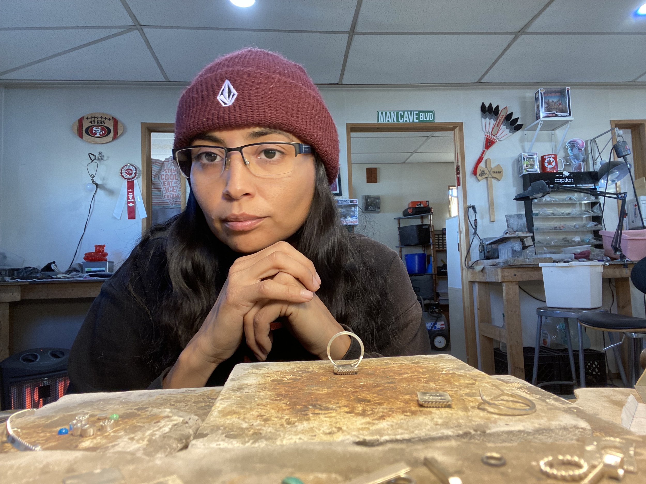 A woman with glasses and a red beanie looks at the camera with her hands clasped in front of her, surrounded by jewelry, in a workshop or craft room.