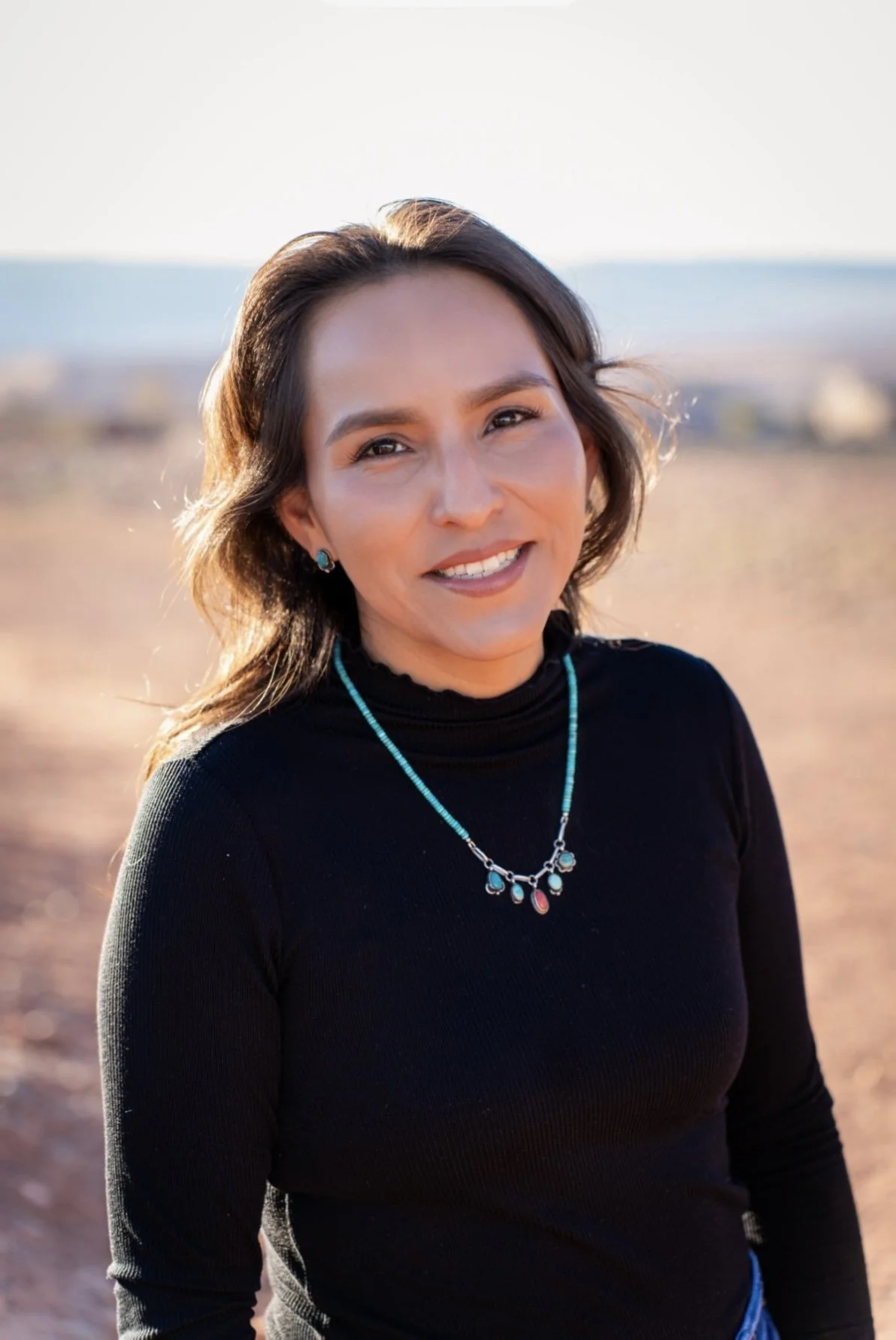 A woman with shoulder-length brown hair smiling outdoors, wearing a black turtleneck sweater and turquoise jewelry, with a blurred desert landscape in the background.