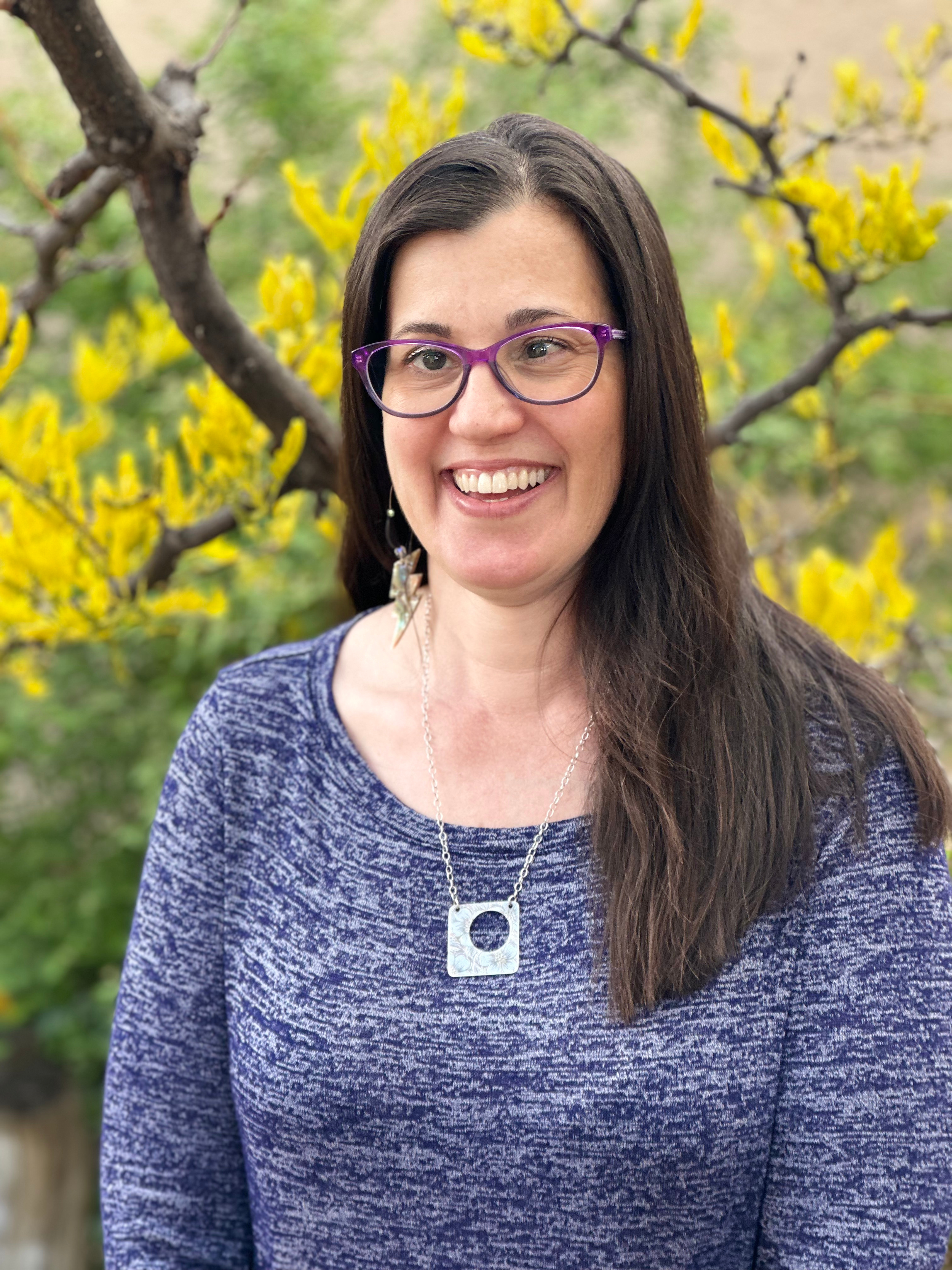 A woman with purple glasses and long dark hair smiling outdoors, standing in front of a tree with yellow flowers.