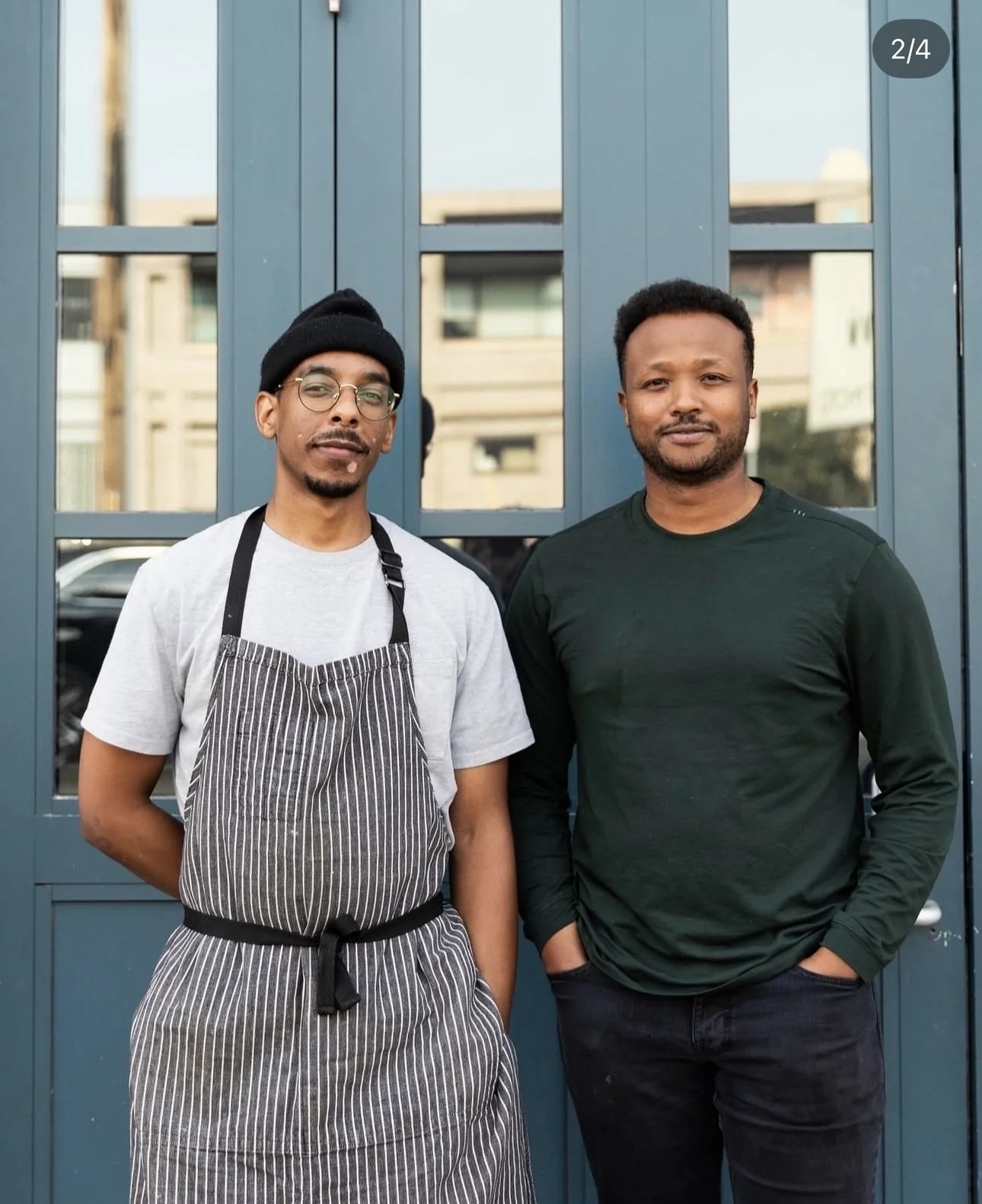 Two men standing in front of a blue door outside a building. The man on the left is wearing glasses, a black beanie, a white t-shirt, and a striped apron, and has his hands behind his back. The man on the right is wearing a dark green long-sleeve shirt and black pants, with his hand in his pocket.