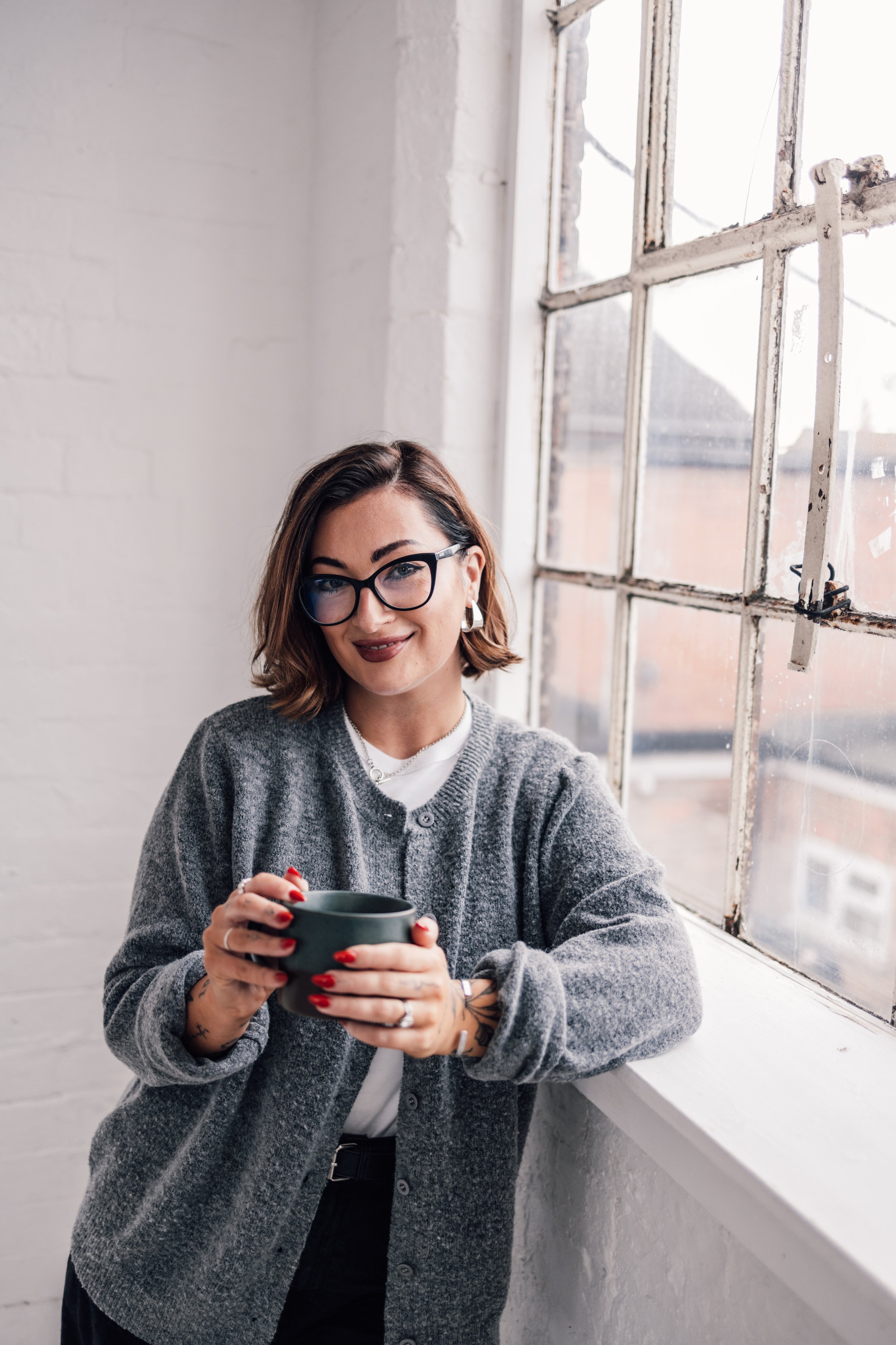 A woman with short brown hair, glasses, and tattoos on her hands holding a dark mug, standing near a large window with white bricks.