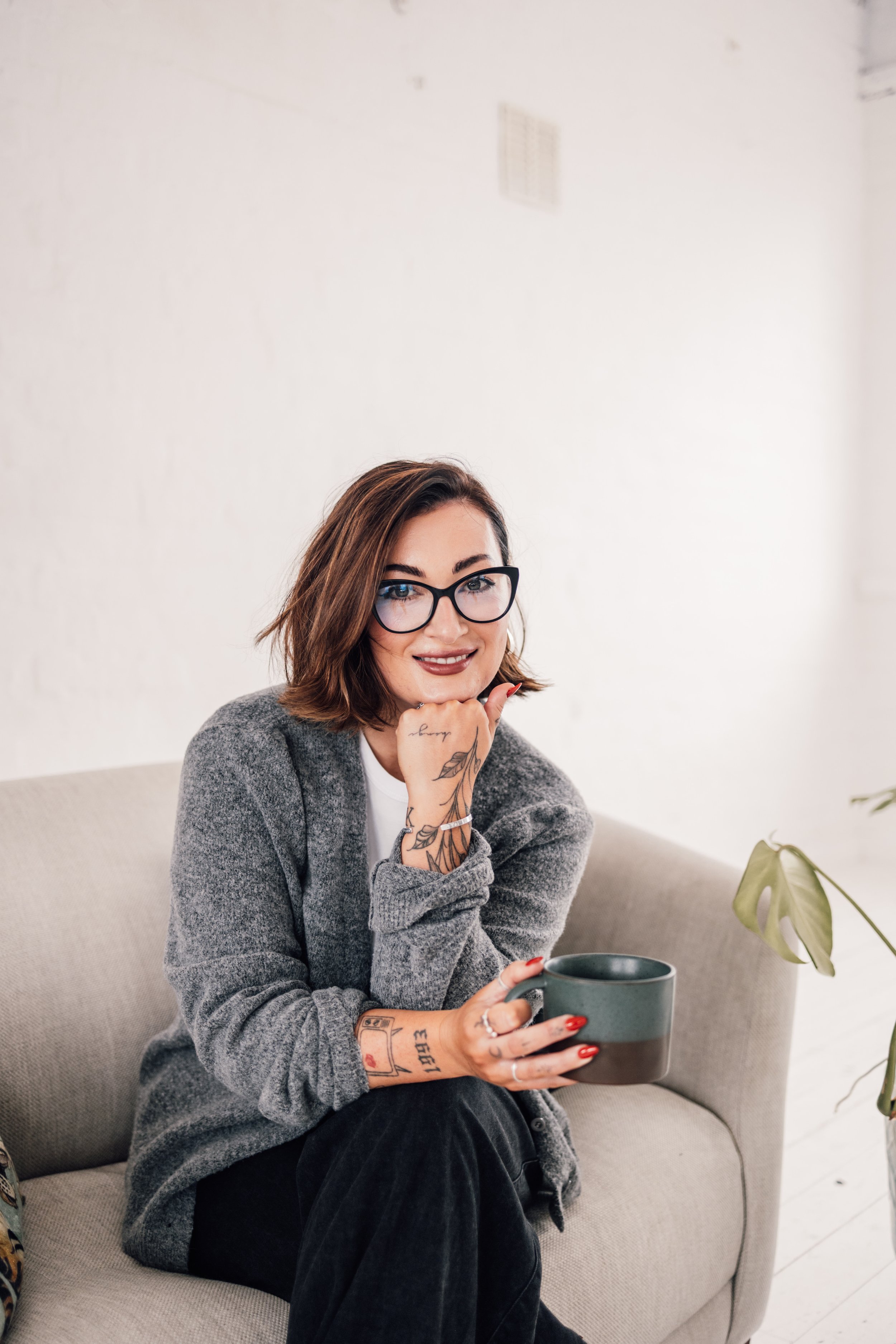 A woman with brown hair, glasses, and tattoos wearing a gray sweater, sitting on a beige couch, holding a mug, smiling, with a plant and minimalistic background.