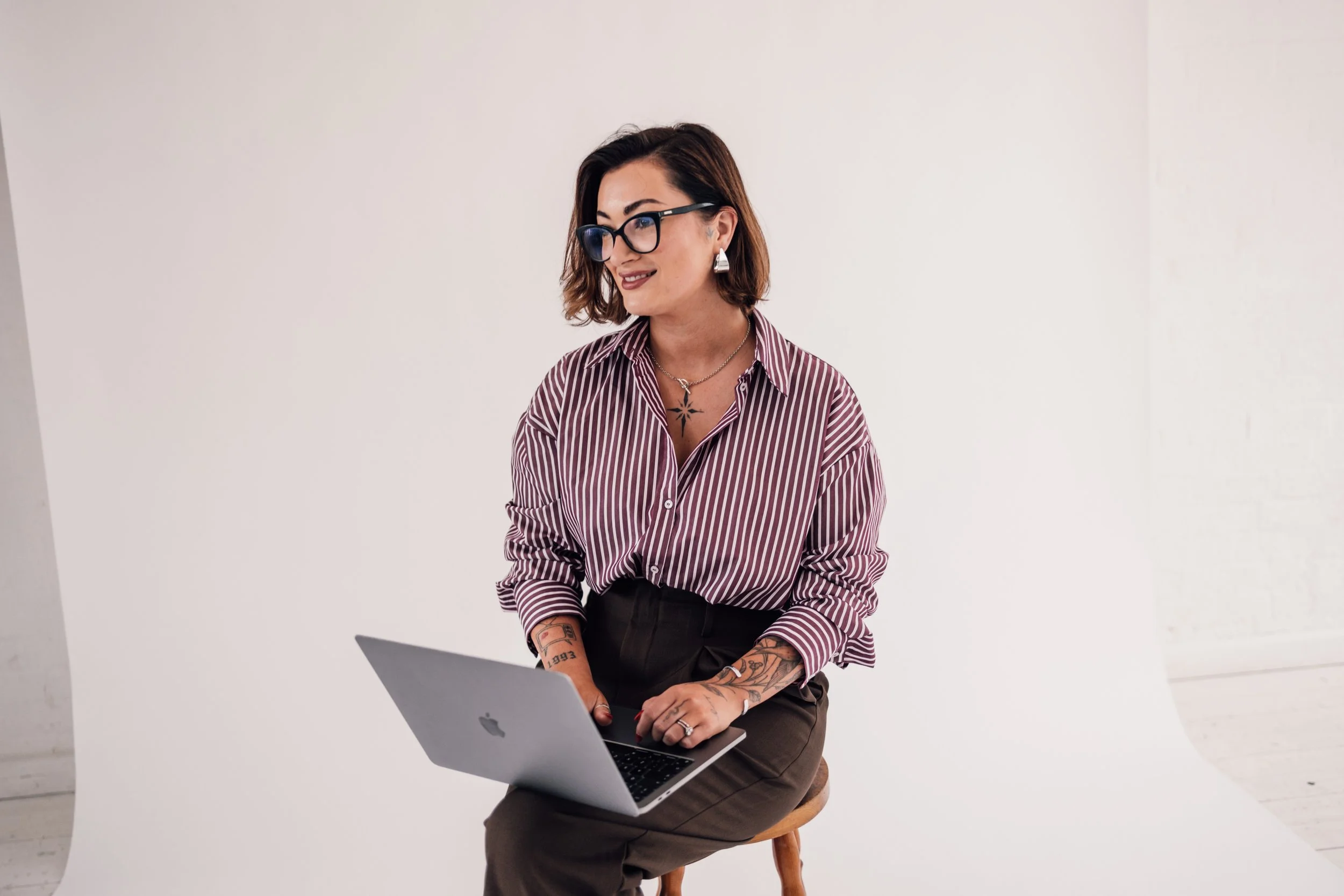 Woman with short brown hair, glasses, and tattoos using a laptop in a minimal studio.