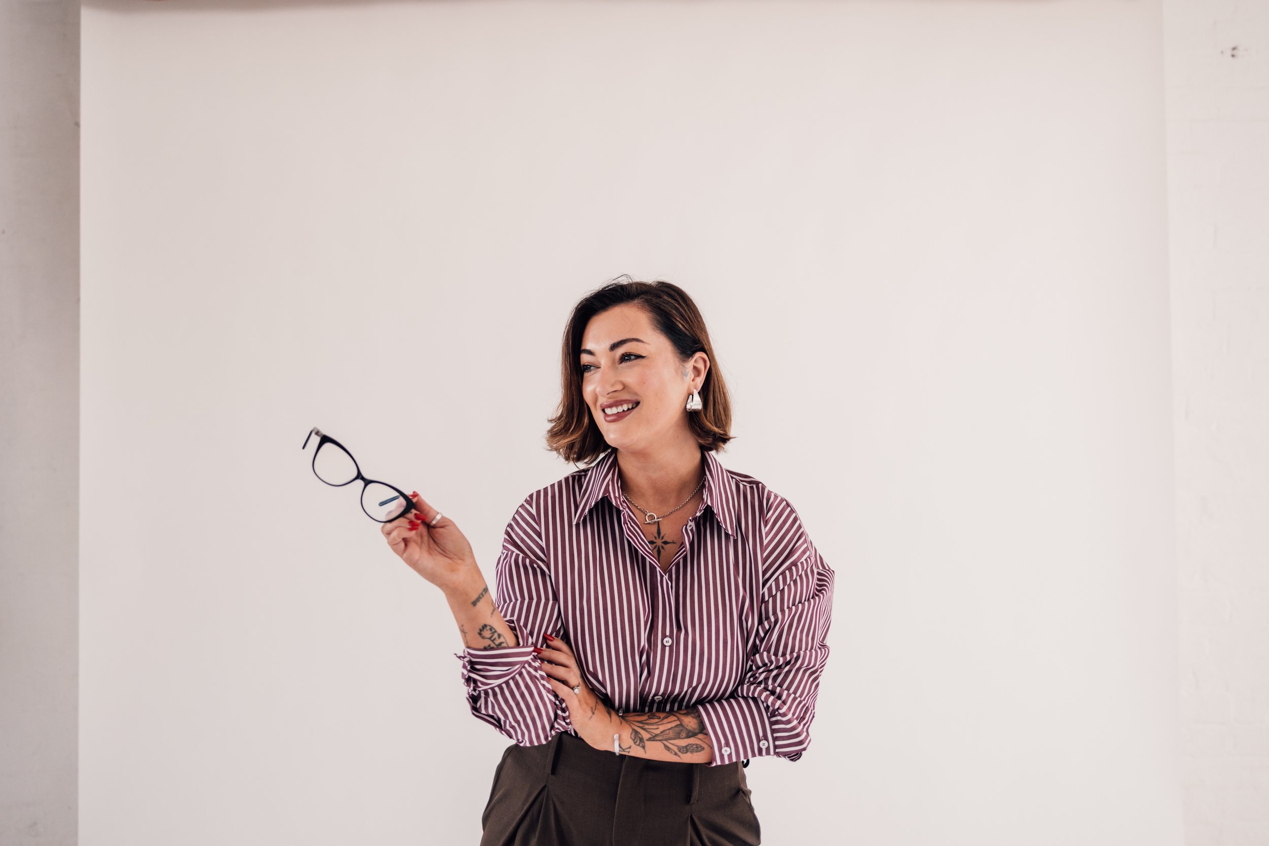 A woman with short brown hair, wearing a striped button-up shirt and dark pants, holding glasses in her right hand, smiling against a plain white background.