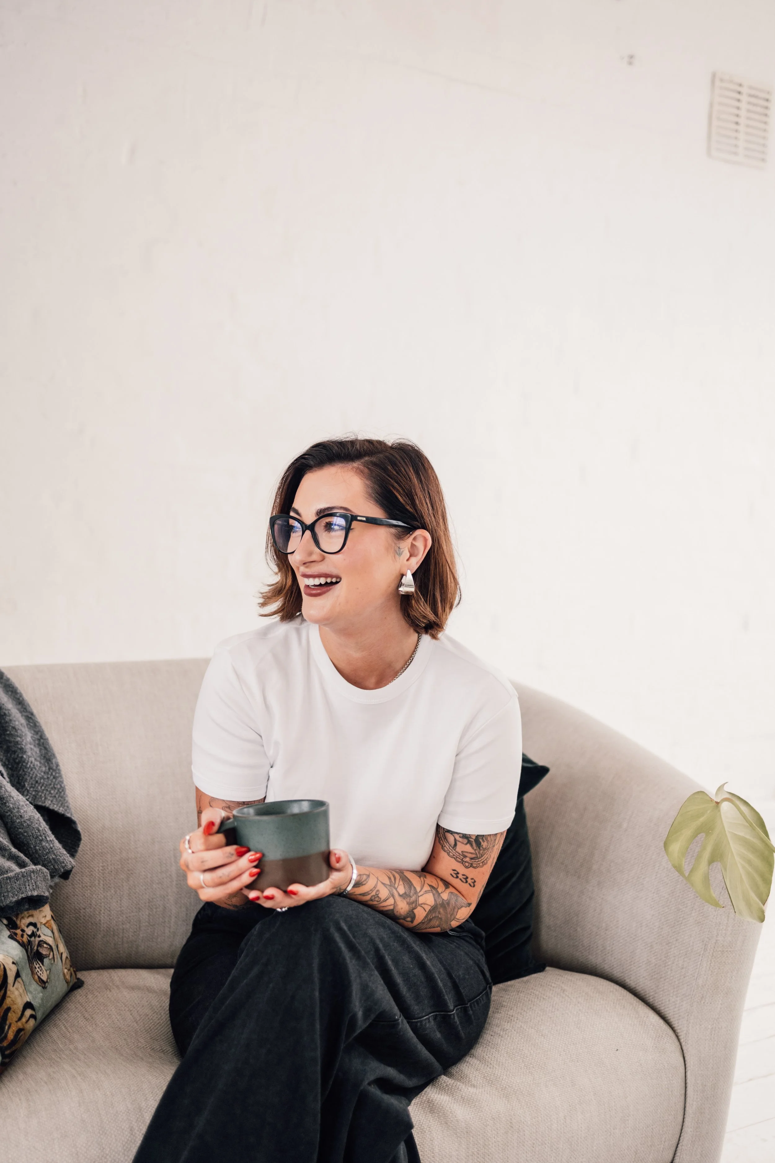 A woman with glasses and tattoos smiling and sitting on a beige couch while holding a dark green mug.