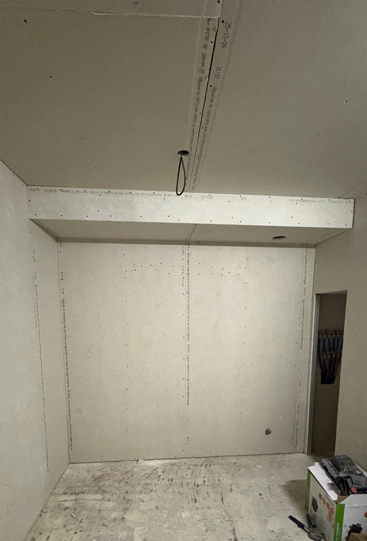Une pièce en construction avec des murs et un plafond en plaques de plâtre, un câble électrique suspendu au plafond, une boîte à outils sur le sol, et une petite ouverture dans le mur à droite.