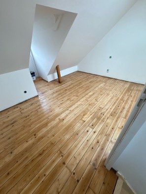 Pièce vide avec parquet en bois clair, murs blancs, dans un grenier ou loft avec une pente de toit et une petite ouverture. Non meublée, prête à être aménagée.