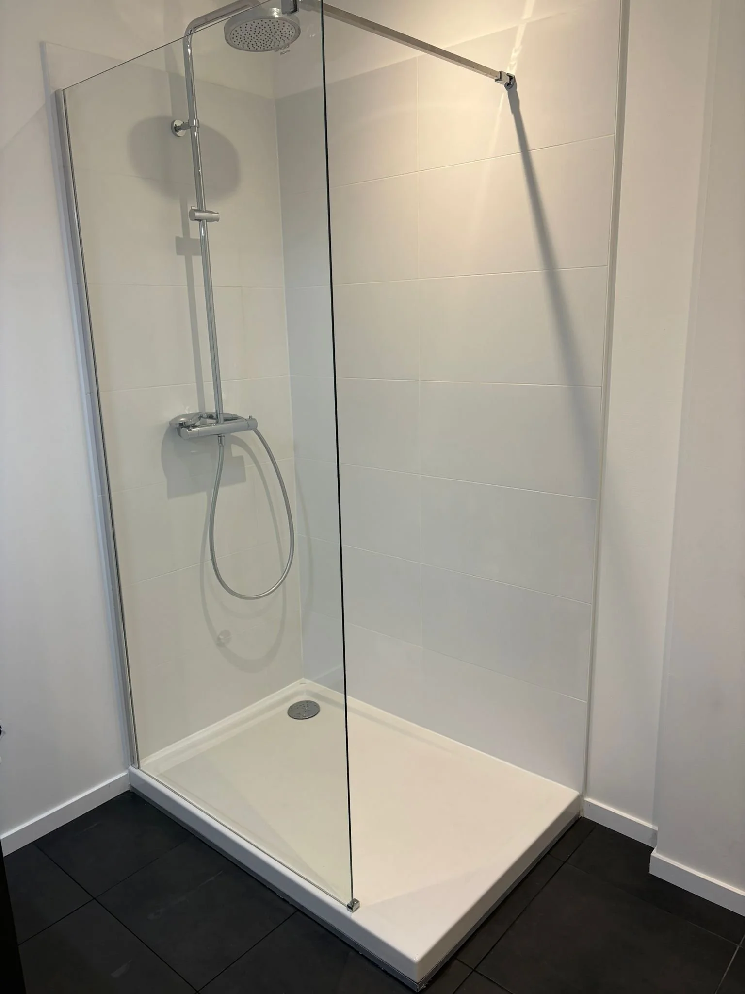 Douche moderne avec paroi en verre, pomme de douche ronde, fiche au plafond, et sol de douche blanc séparé du sol noir des toilettes.