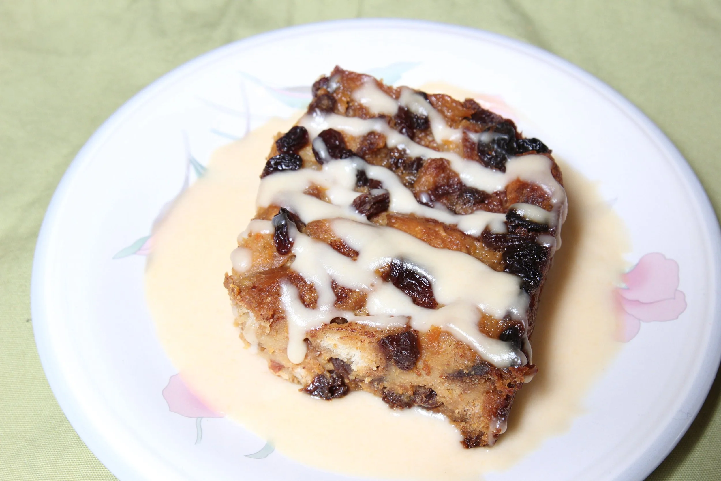 A slice of bread pudding with raisins, drizzled with white icing, served on a white plate with a pink floral pattern, on a green tablecloth.