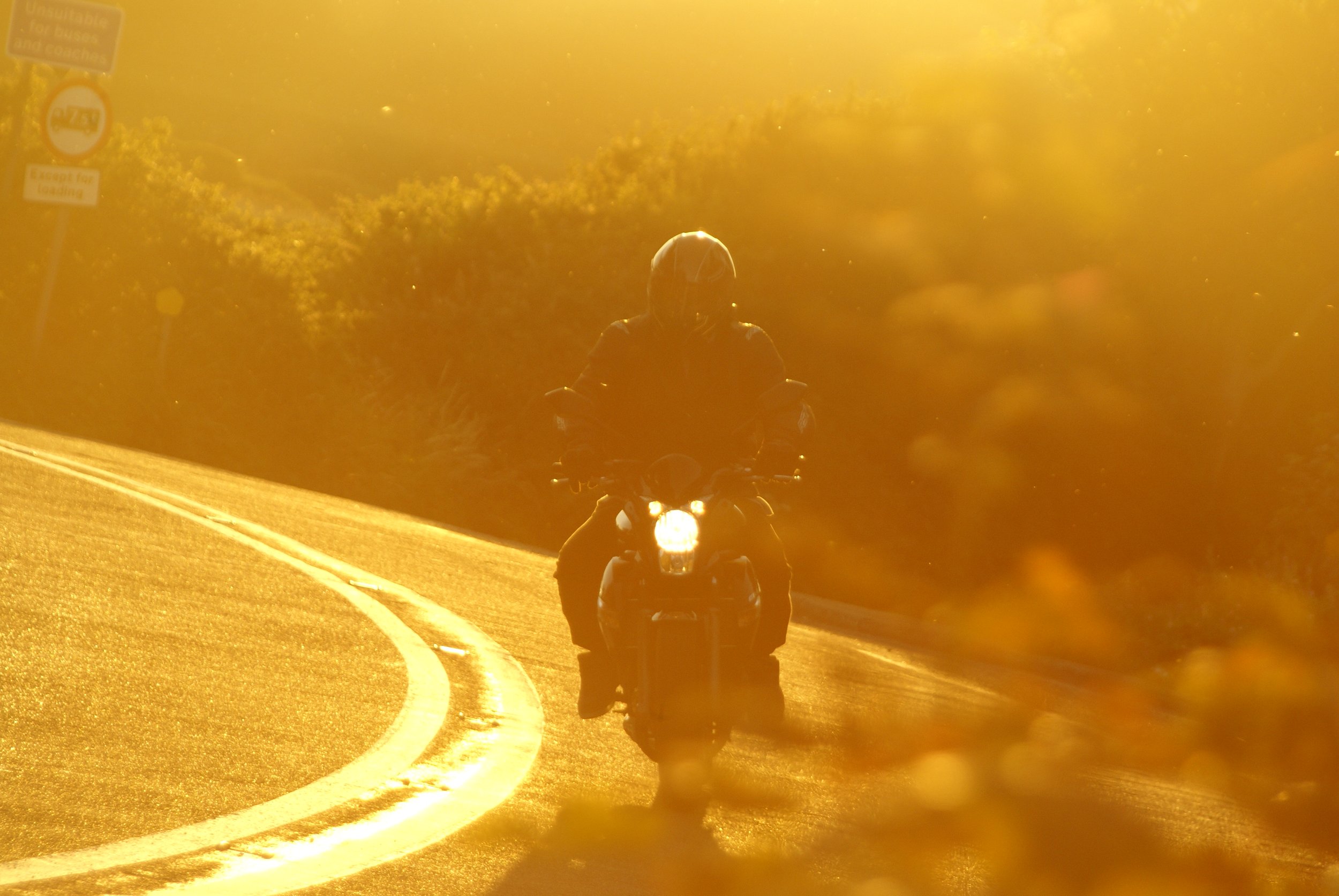 A person riding a motorcycle on a curved road during sunset or sunrise, with a bright yellow-orange hue illuminating the scene.