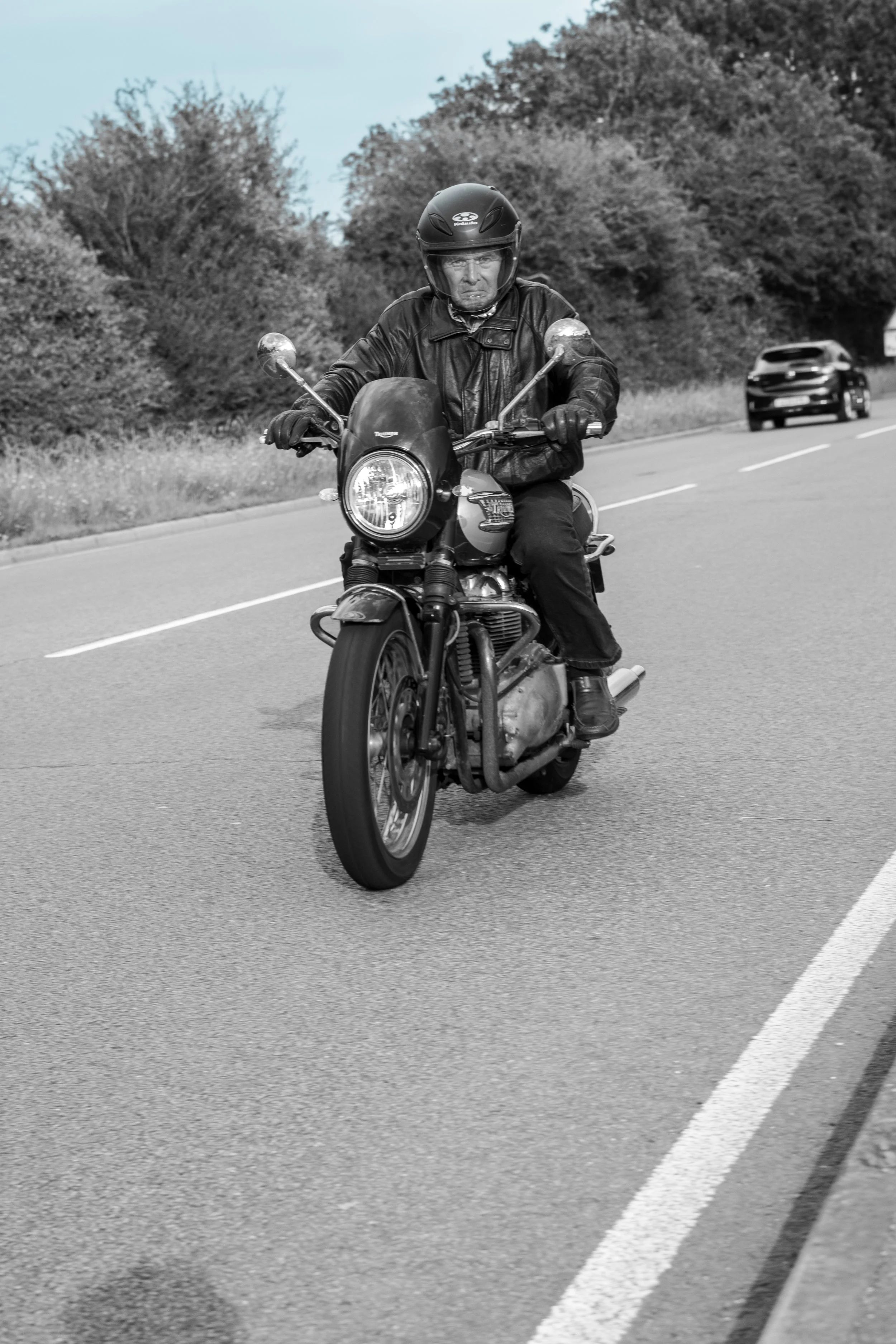 A man wearing a black helmet and leather jacket riding a motorcycle on a two-lane road with other vehicles and trees in the background.