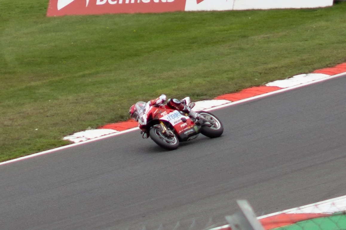 Motorcycle racer in red gear leaning into a turn on a race track with a grassy area and advertising boards in the background.