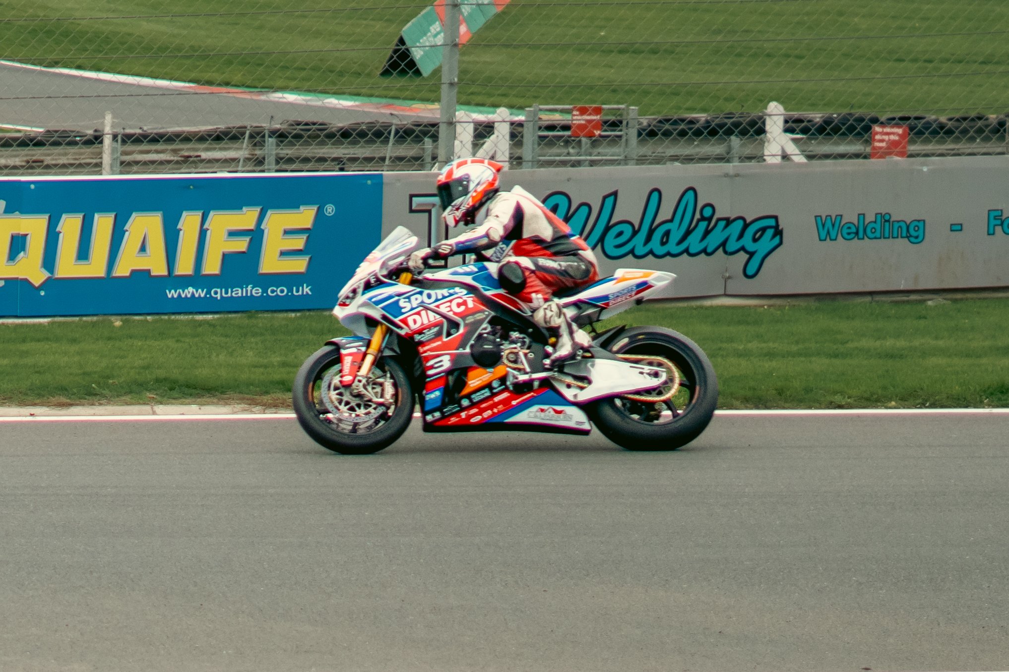 Motorsport rider in white and red racing suit on a colorful motorcycle racing on a track, with advertising banners in the background.
