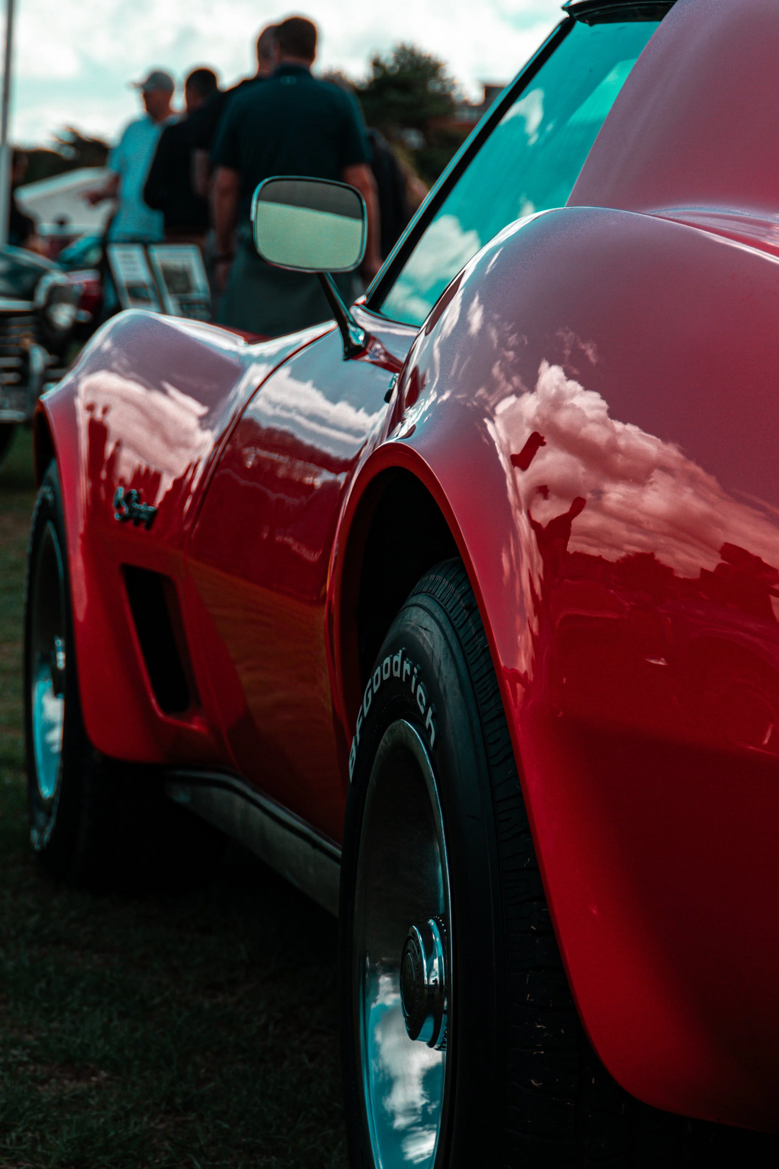 Close-up of a red sports car with a reflective surface showing blue sky and clouds, at an outdoor event with people in the background.