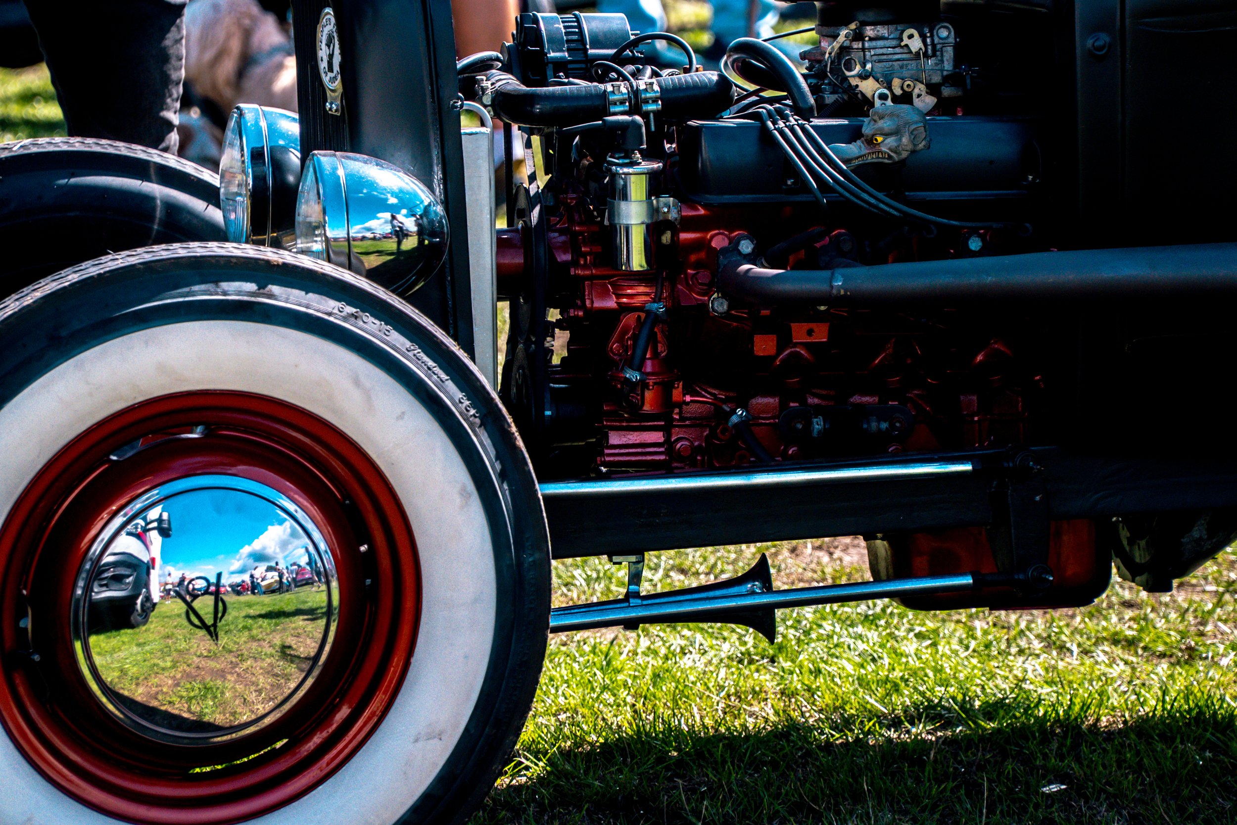 Close-up of a vintage car engine with a shiny headlamp and whitewall tire, reflecting the blue sky and clouds, with grass on the ground and people in the background.