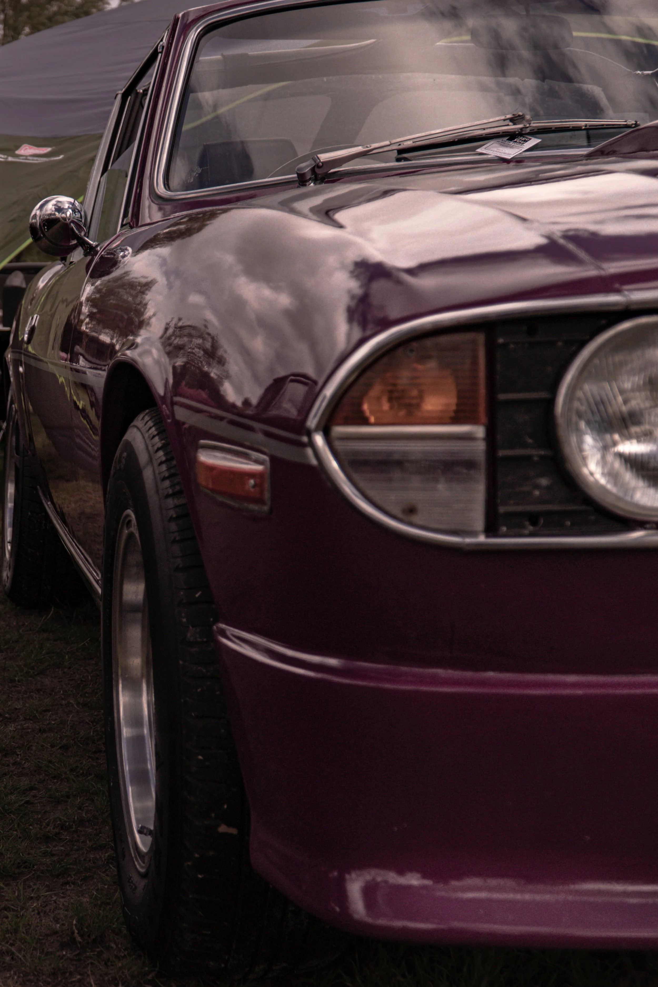 Close-up of the front of a vintage burgundy car, showing the headlight, front fender, and windshield with reflections of cloudy sky.