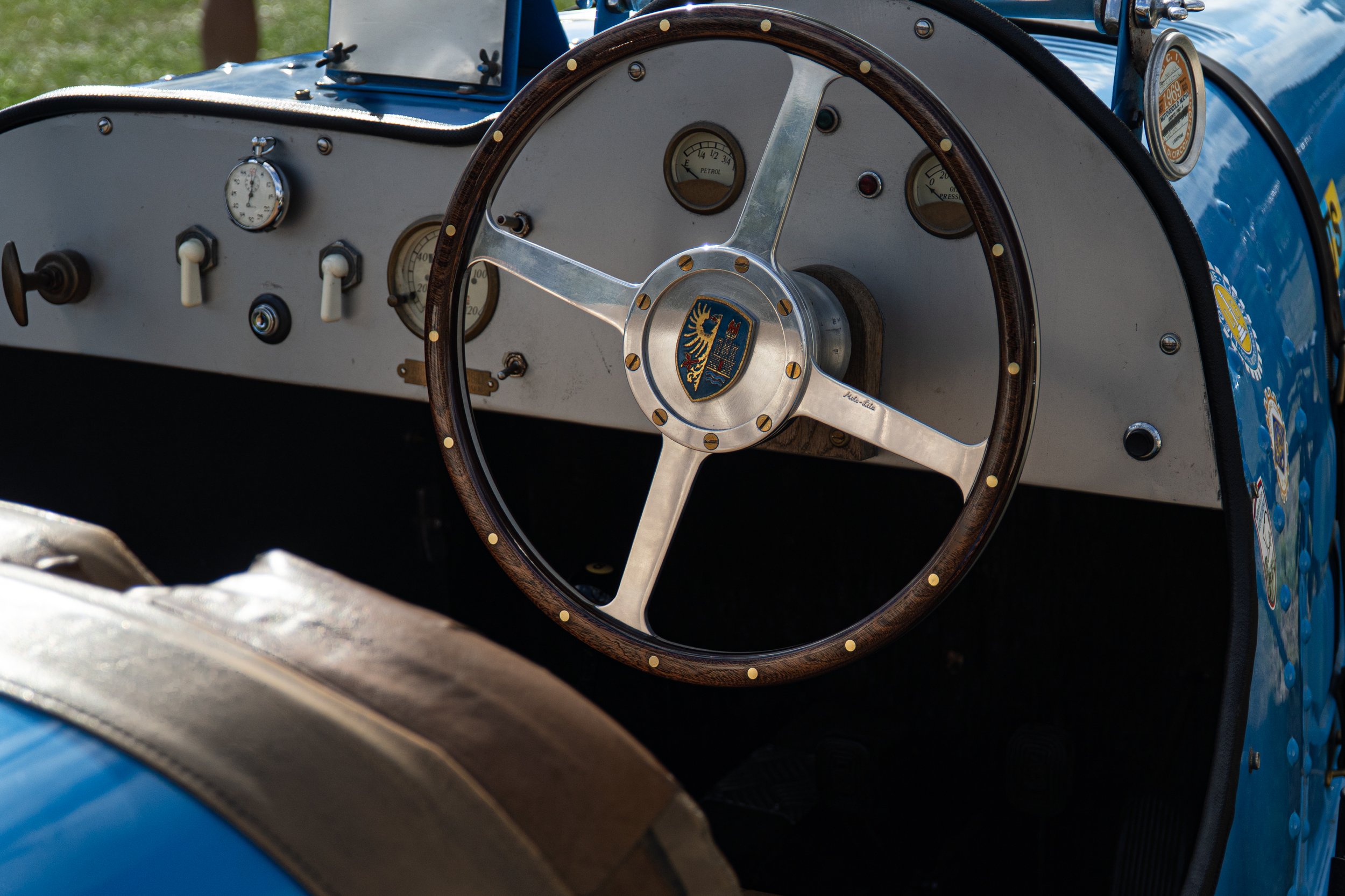 Close-up of the dashboard and steering wheel of a vintage blue race car, showing gauges and a wooden steering wheel with a crest in the center.