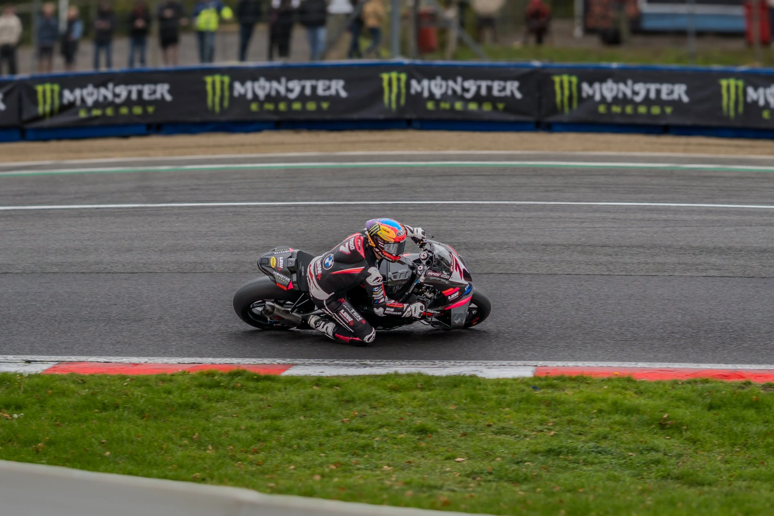 A motorcycle rider in racing gear and a helmet leans into a turn on a race track, with a black barrier displaying 'Monster Energy' logos in the background.