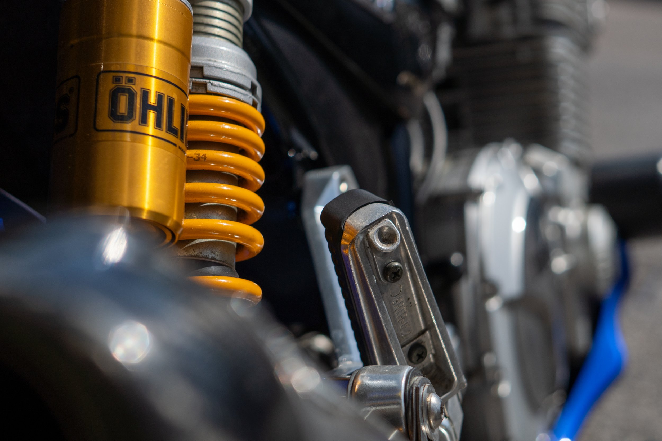 Close-up of motorcycle suspension with a yellow shock absorber, part of the motorcycle's frame and engine components visible, featuring metallic and black parts.