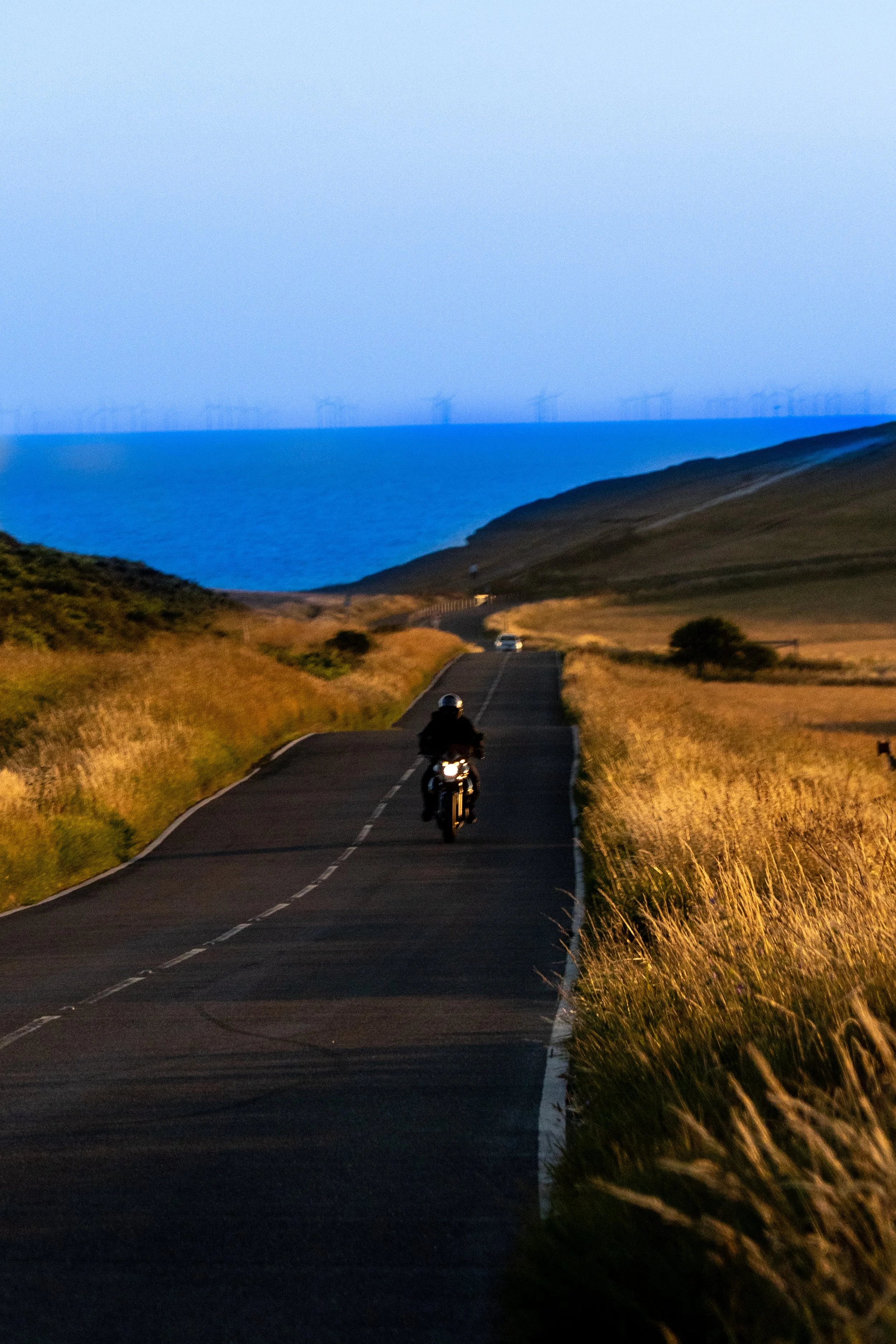 A motorcyclist riding down a long, winding road through green hills during sunset, with wind turbines visible on the horizon over the blue sea.
