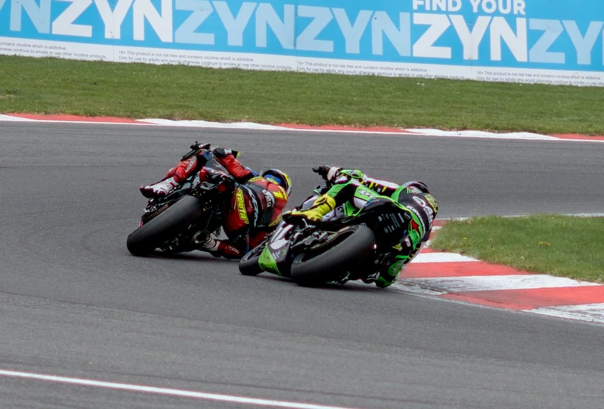 Two motorcycle racers leaning into a sharp turn on a racetrack, with a grass border and a large advertisement banner in the background.
