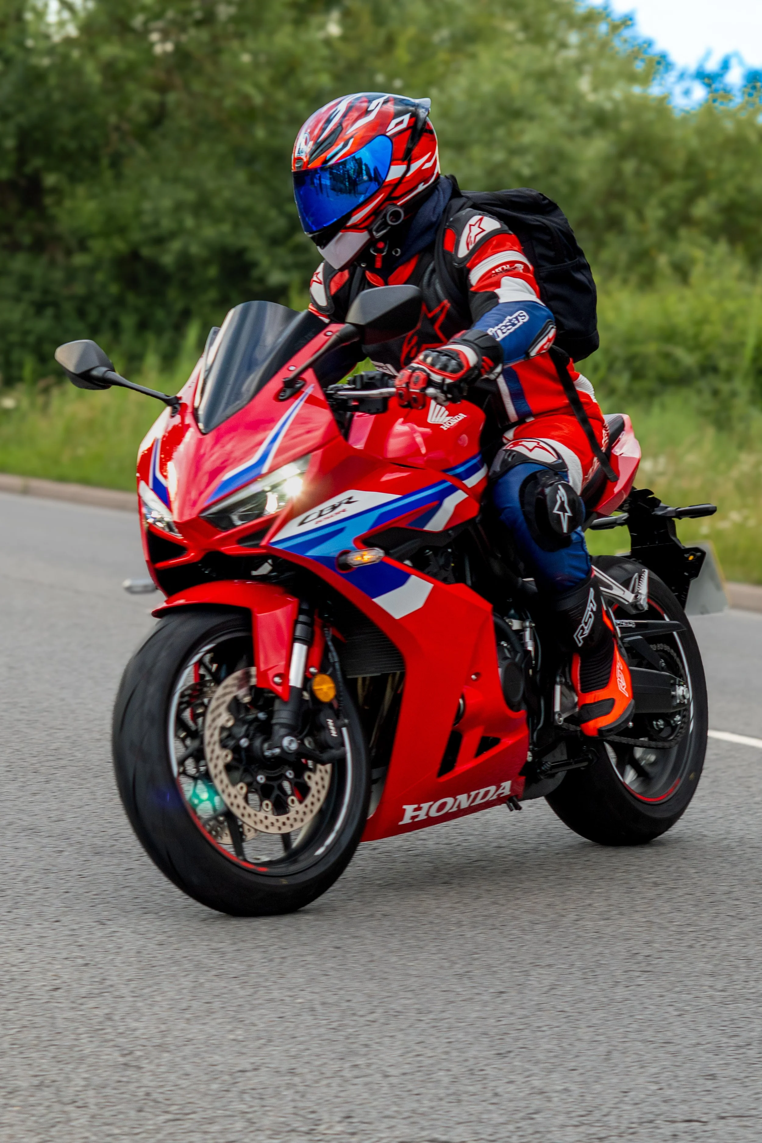 A person wearing a red, black, and blue motorcycle racing suit and helmet riding a red Honda sport motorcycle on a paved road with foliage in the background.