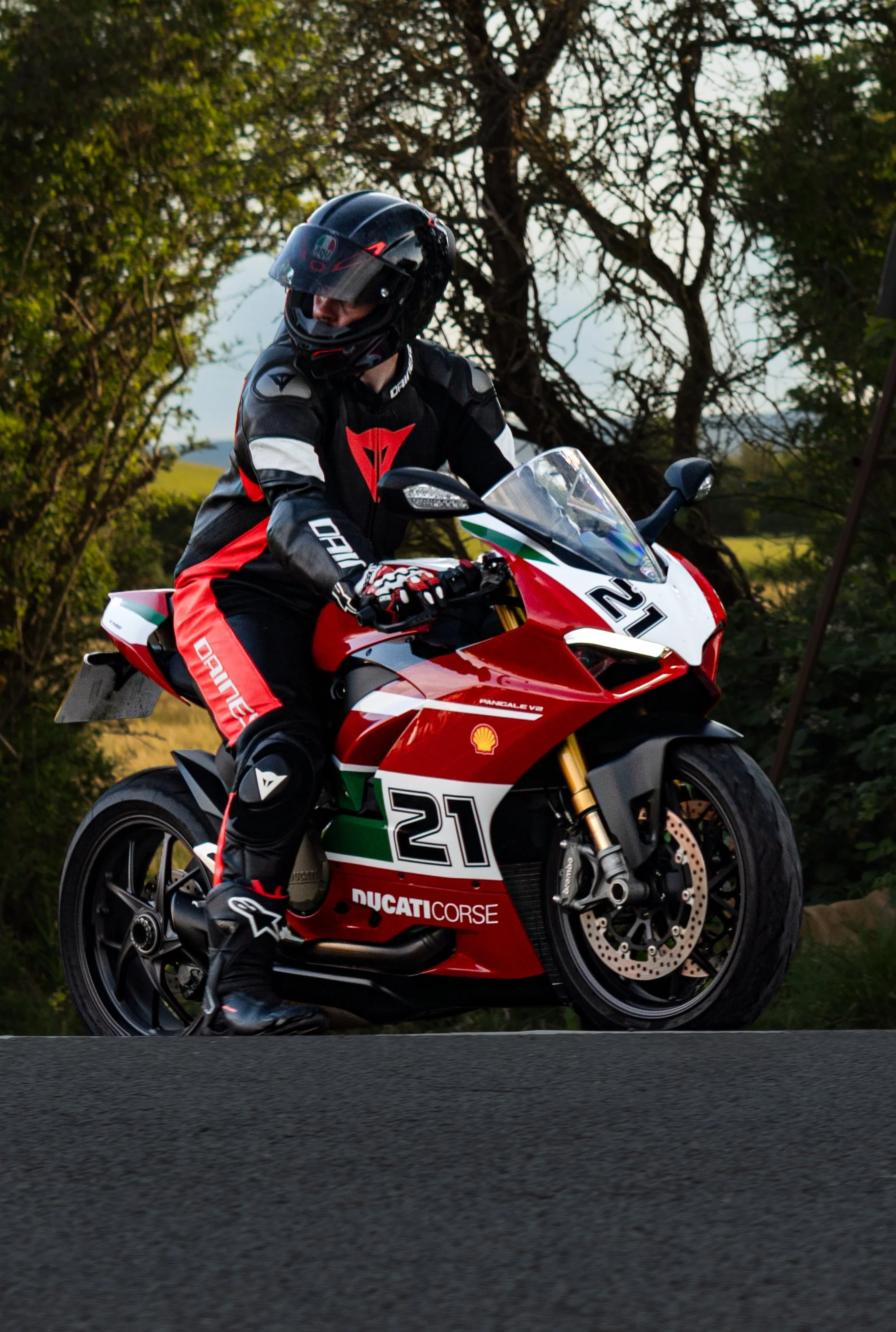 A motorcyclist dressed in racing gear, including a black helmet with a clear visor, a black and red racing suit with white accents, riding a red Ducati motorcycle on an outdoor road with trees in the background.