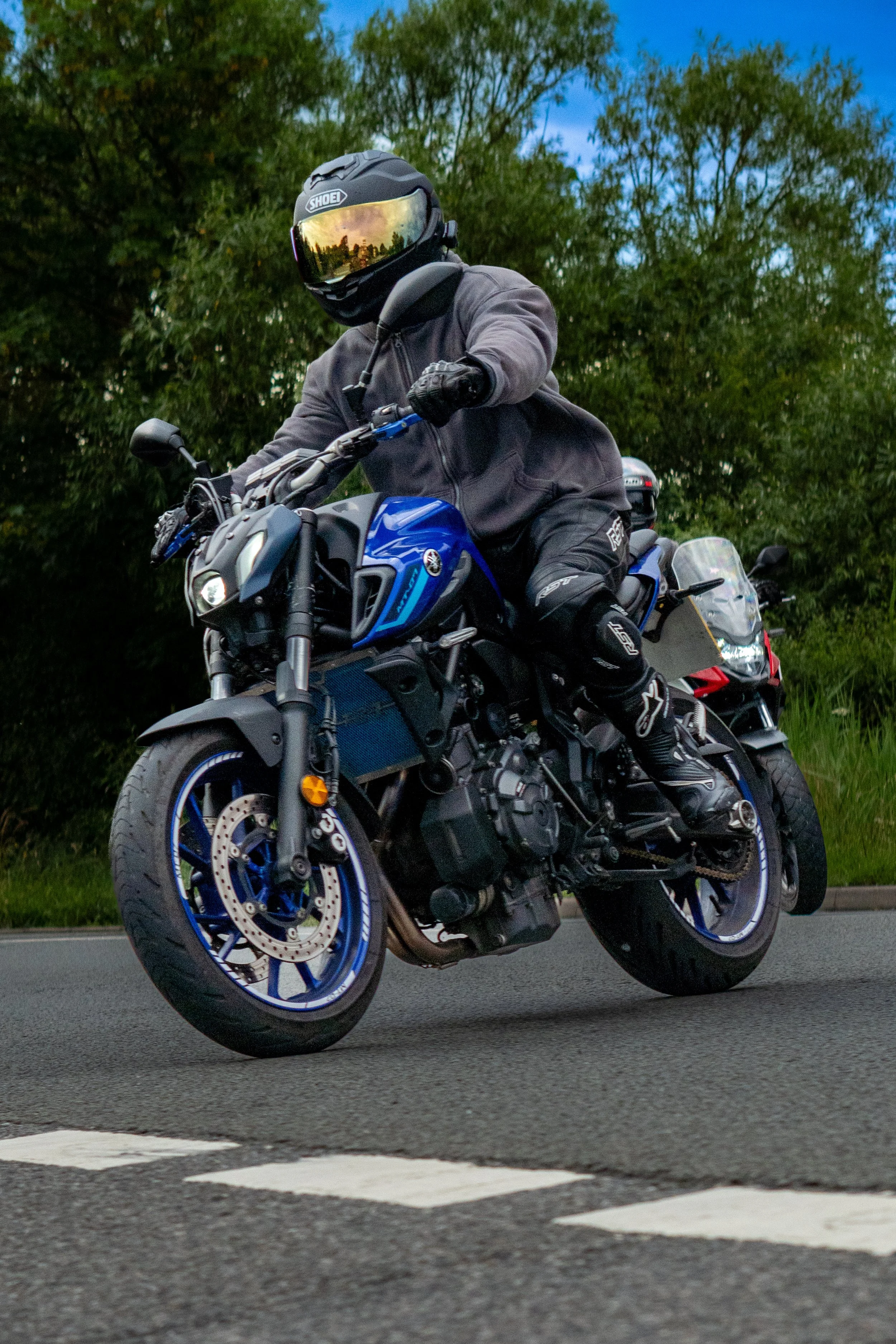 Motorcyclist wearing a black helmet with a reflective visor, gray jacket, and black gloves riding a blue Yamaha motorcycle on a paved road with green trees in the background.