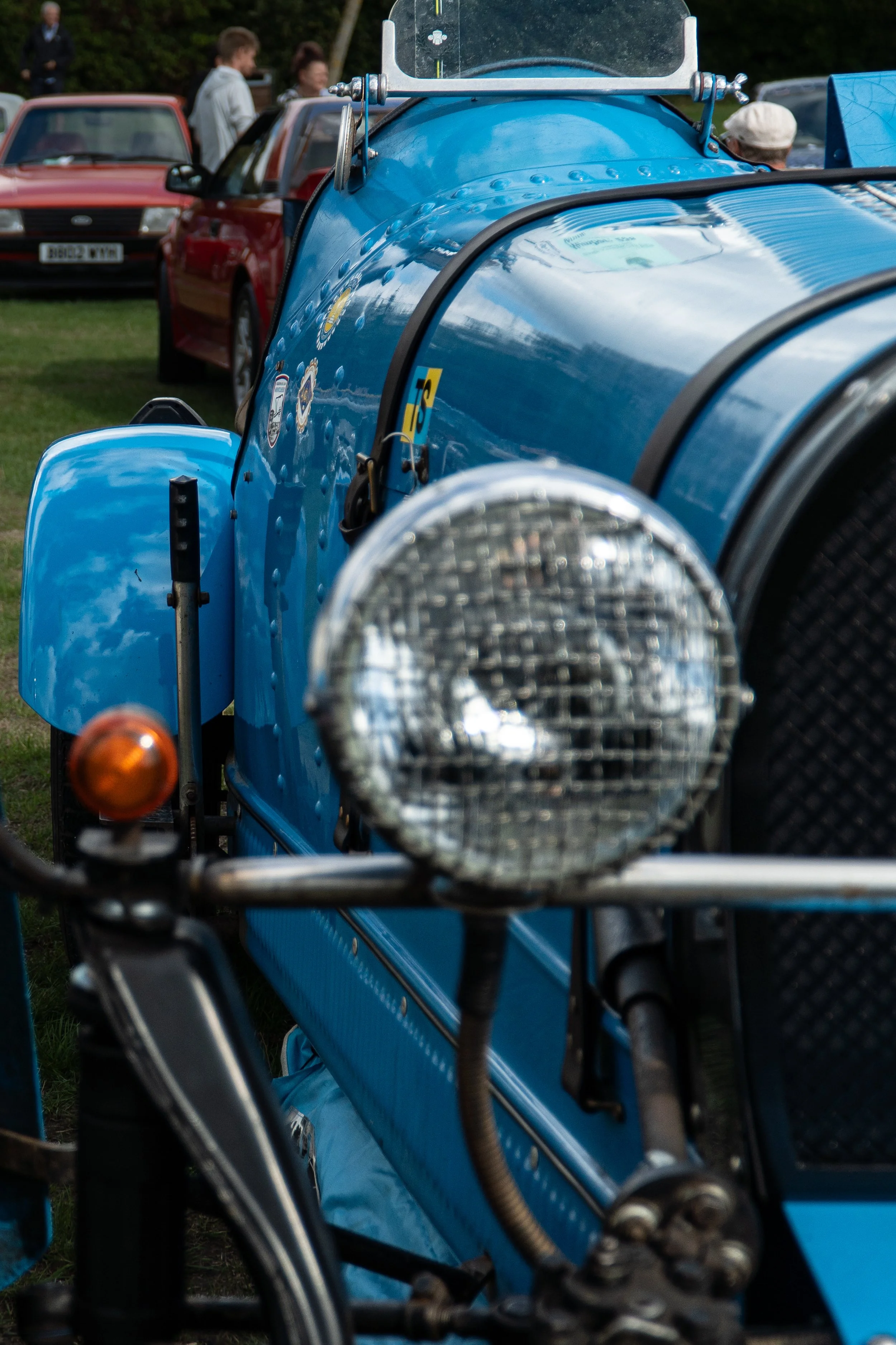 Close-up of a vintage blue race car with its front headlight and bumper visible, parked at a car show with people and other cars in the background.