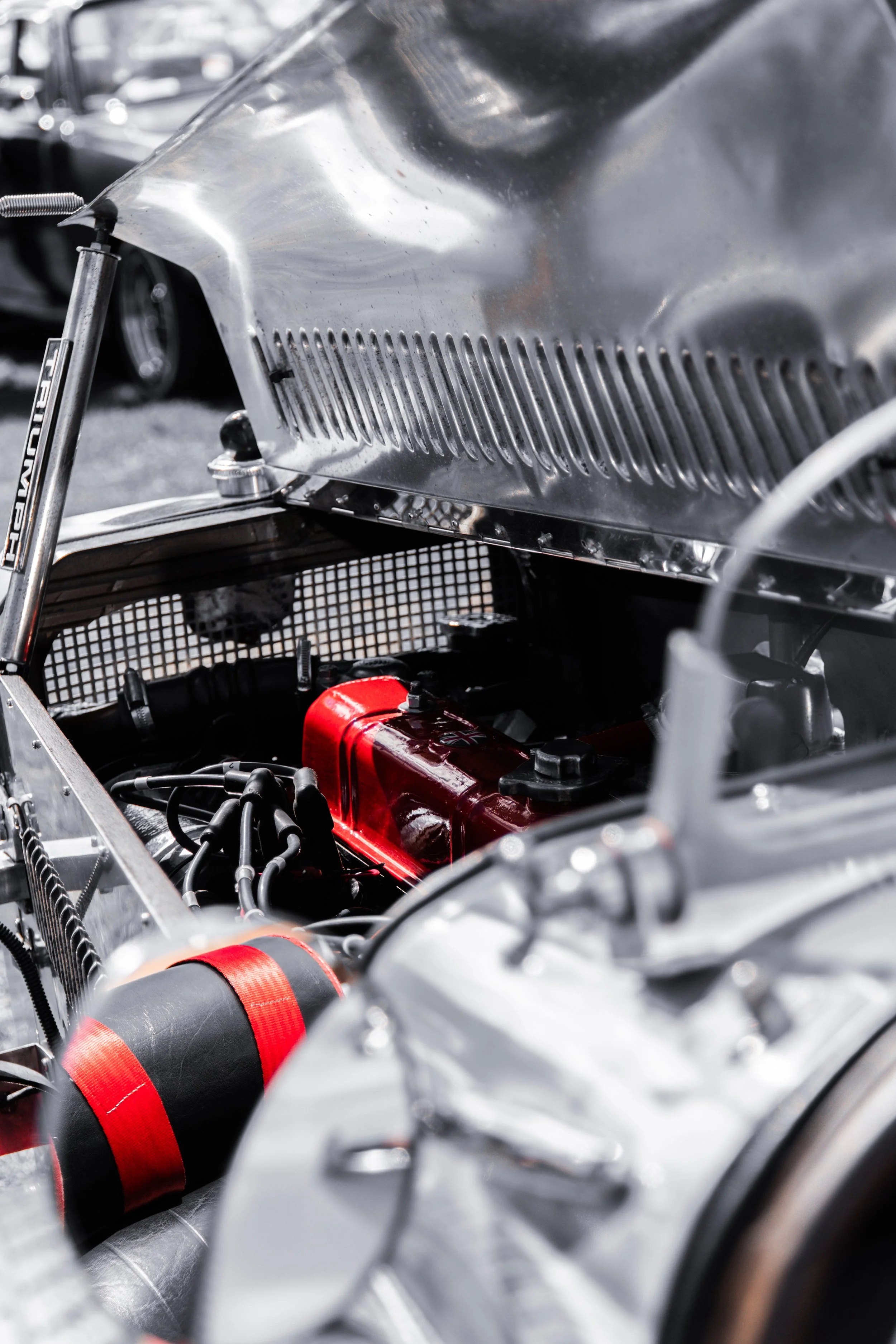 Close-up of a vintage racing car engine, displaying metallic parts, red accents on hoses, and detailed mechanical components.